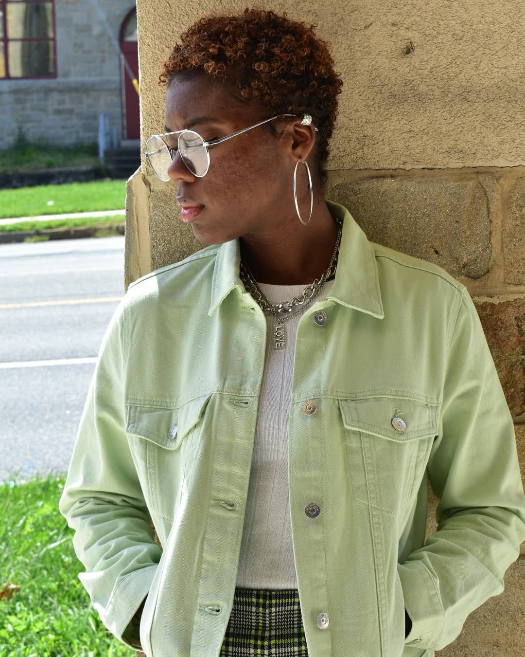 Young Black woman in mint green denim jacket and aviator sunglasses poses against stone wall outdoors.