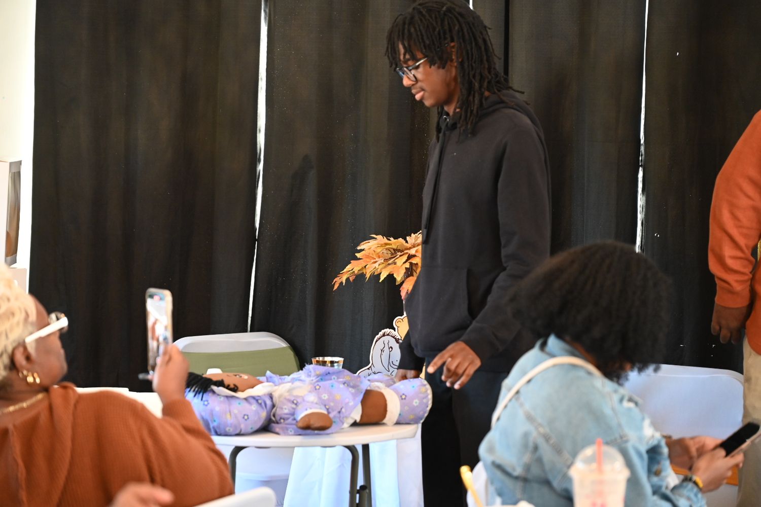 A tall man with dreadlocks stands at a baby shower table examining items near a baby doll display.