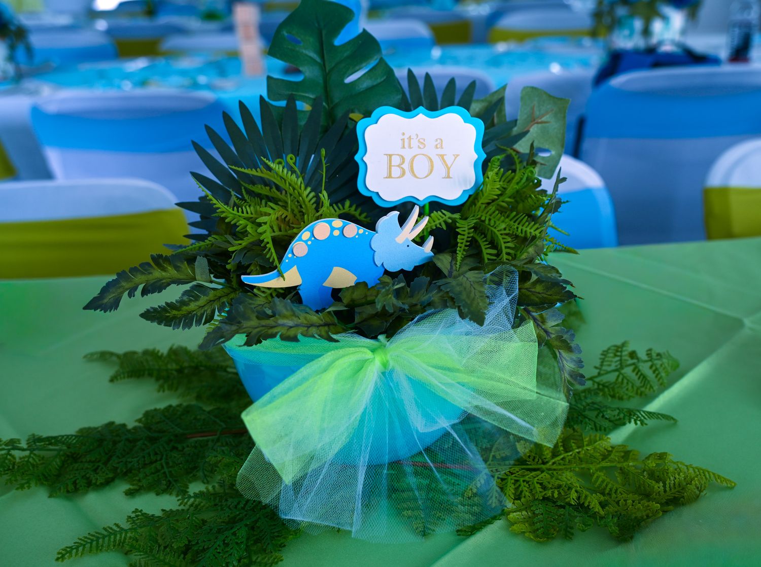 Dinosaur-themed baby boy shower centerpiece with ferns, triceratops figurine, and green tulle bow.