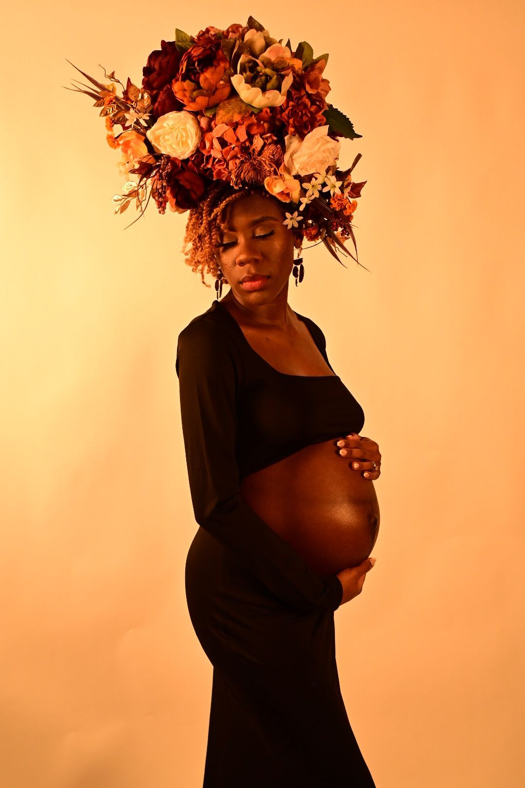 Pregnant Black woman in black dress with eyes closed, wearing autumn floral crown, holding belly.