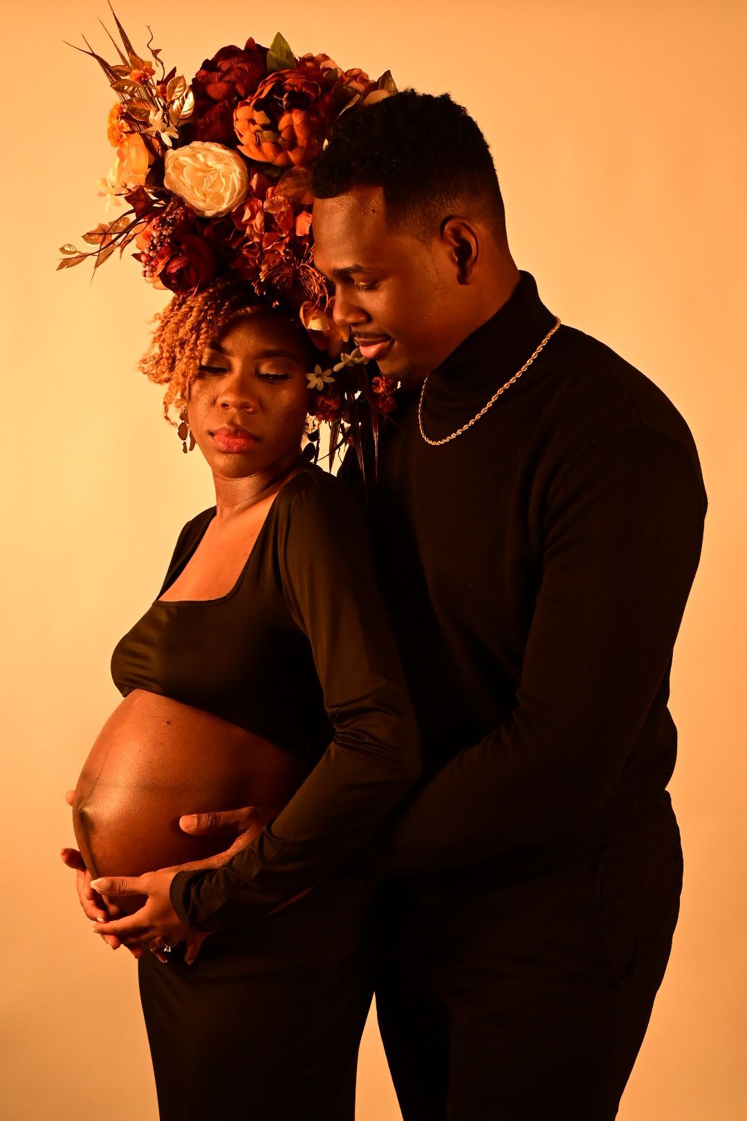 Couple embracing during maternity photoshoot, pregnant woman in floral headdress and black outfit on golden background.