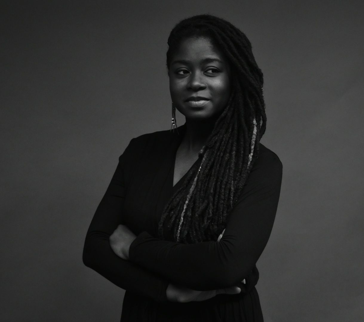 Black and white portrait of a young woman with locs wearing a black top, arms crossed confidently.