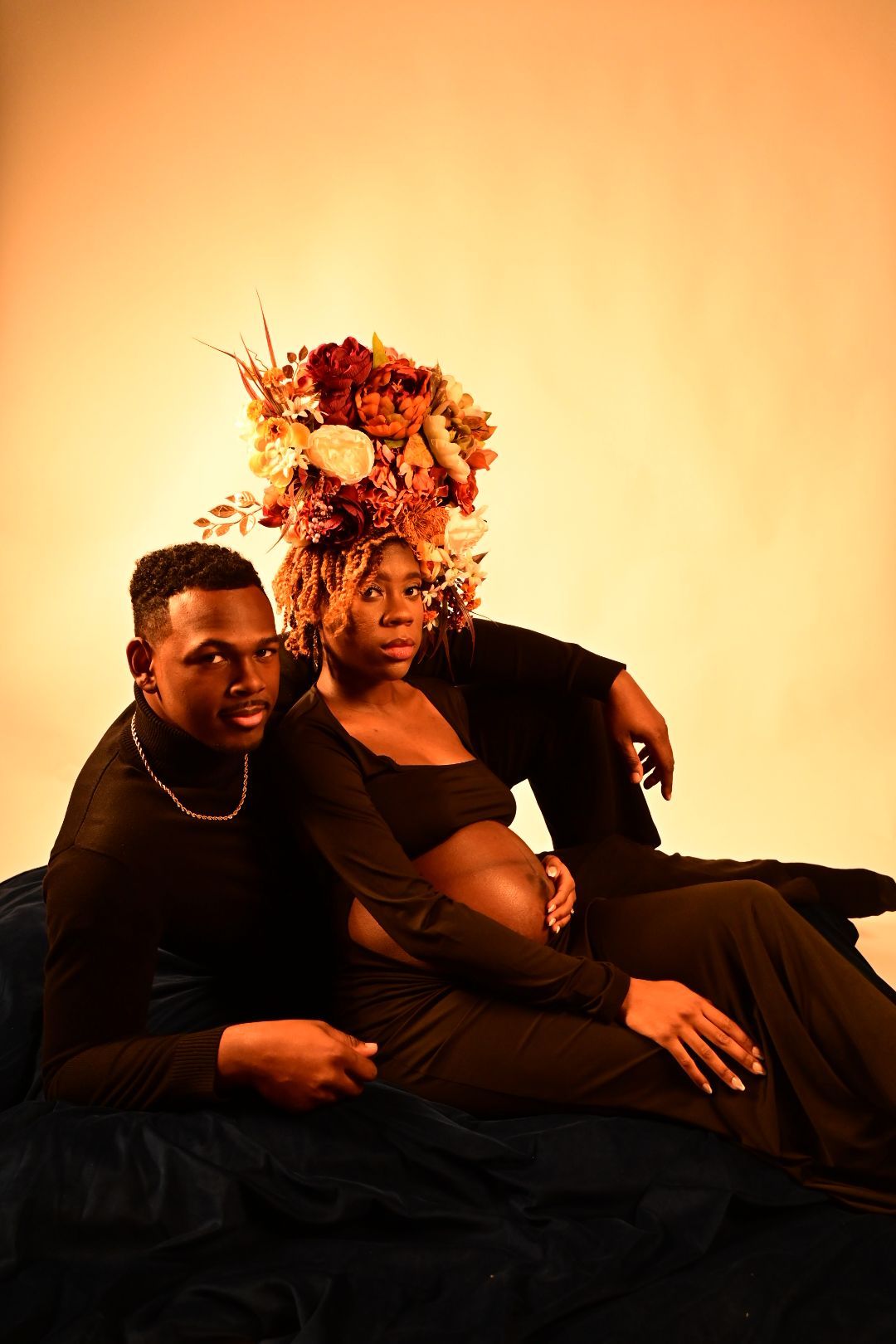 Couple posing for maternity shoot on dark fabric, woman in floral crown and black outfit on warm golden backdrop.