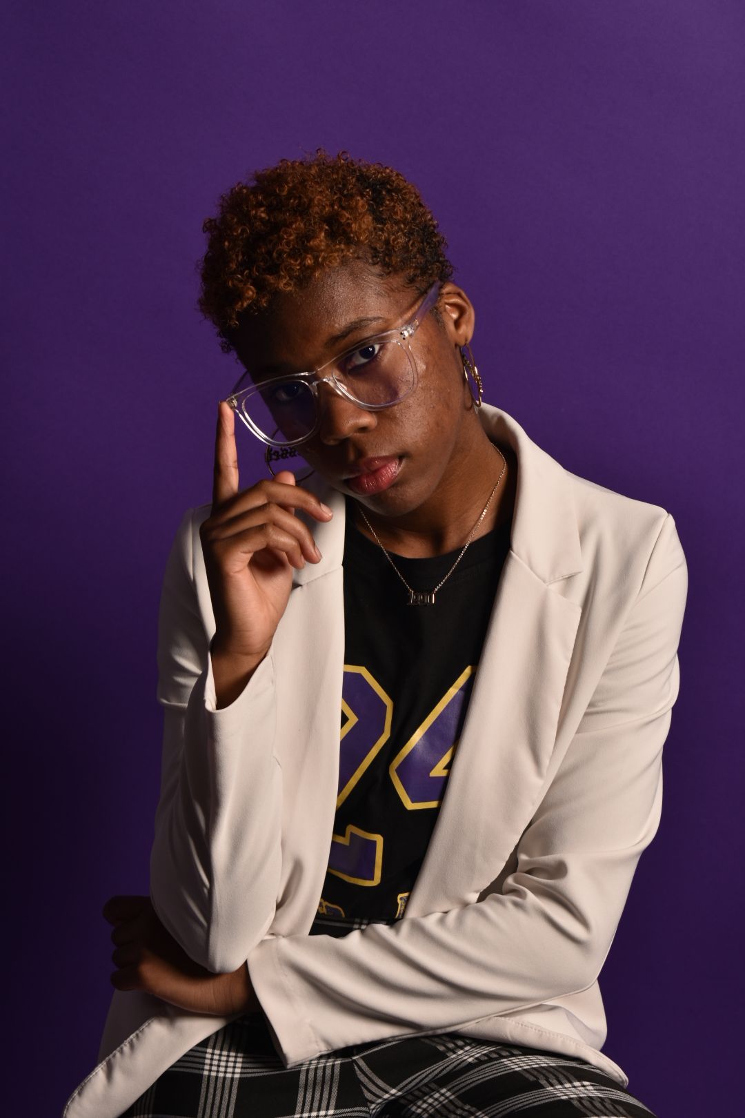 Stylish woman in white blazer and number 24 jersey poses thoughtfully against vibrant purple background.