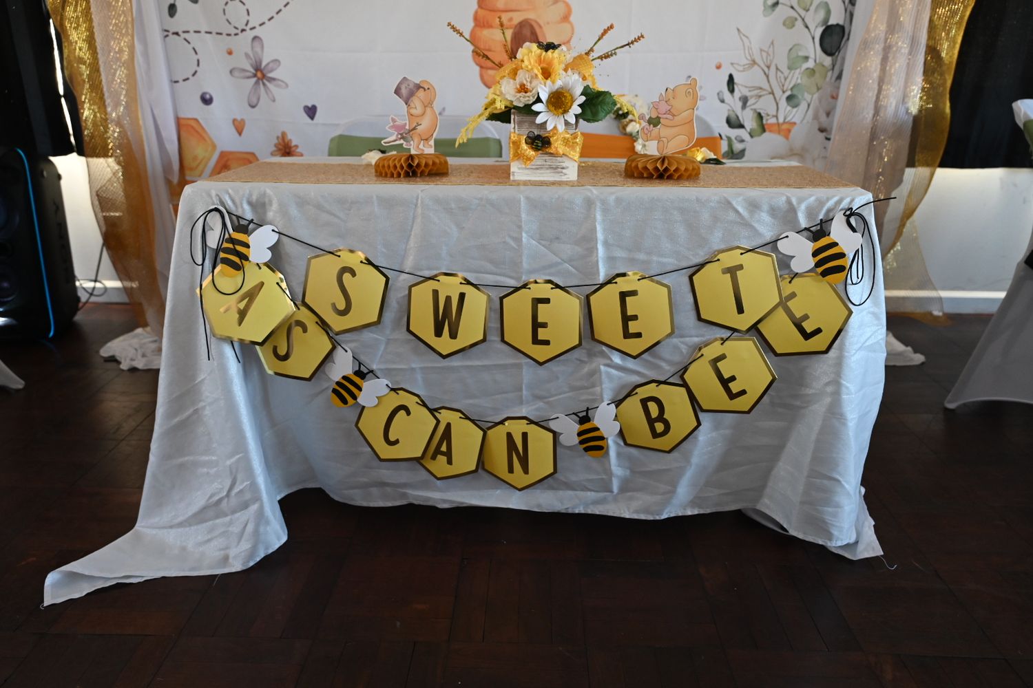 Yellow and black honeycomb banner spelling Sweet As Can Bee decorates a white draped baby shower table.