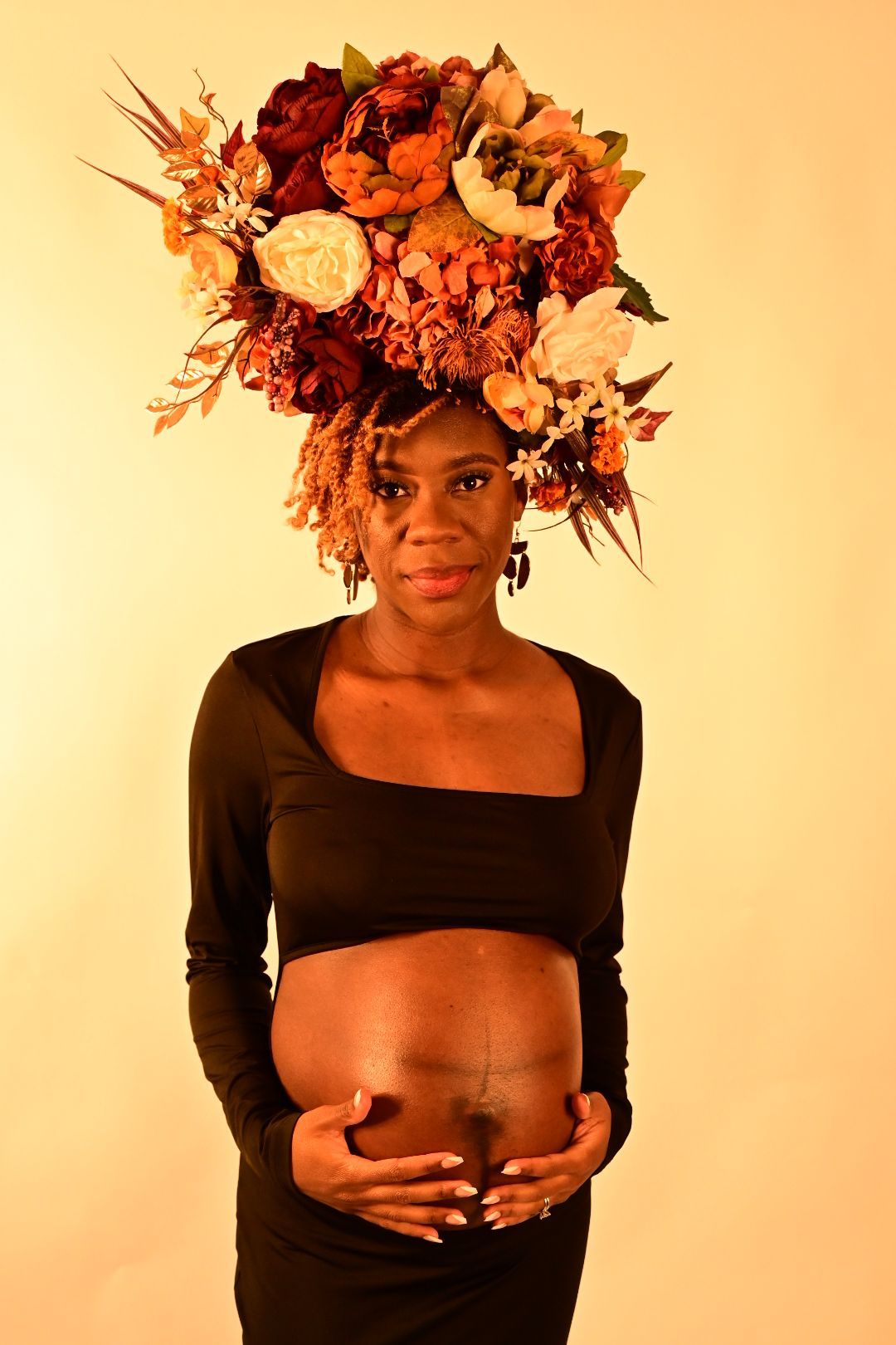 Pregnant Black woman in black crop outfit wearing large floral crown, cradling baby bump.