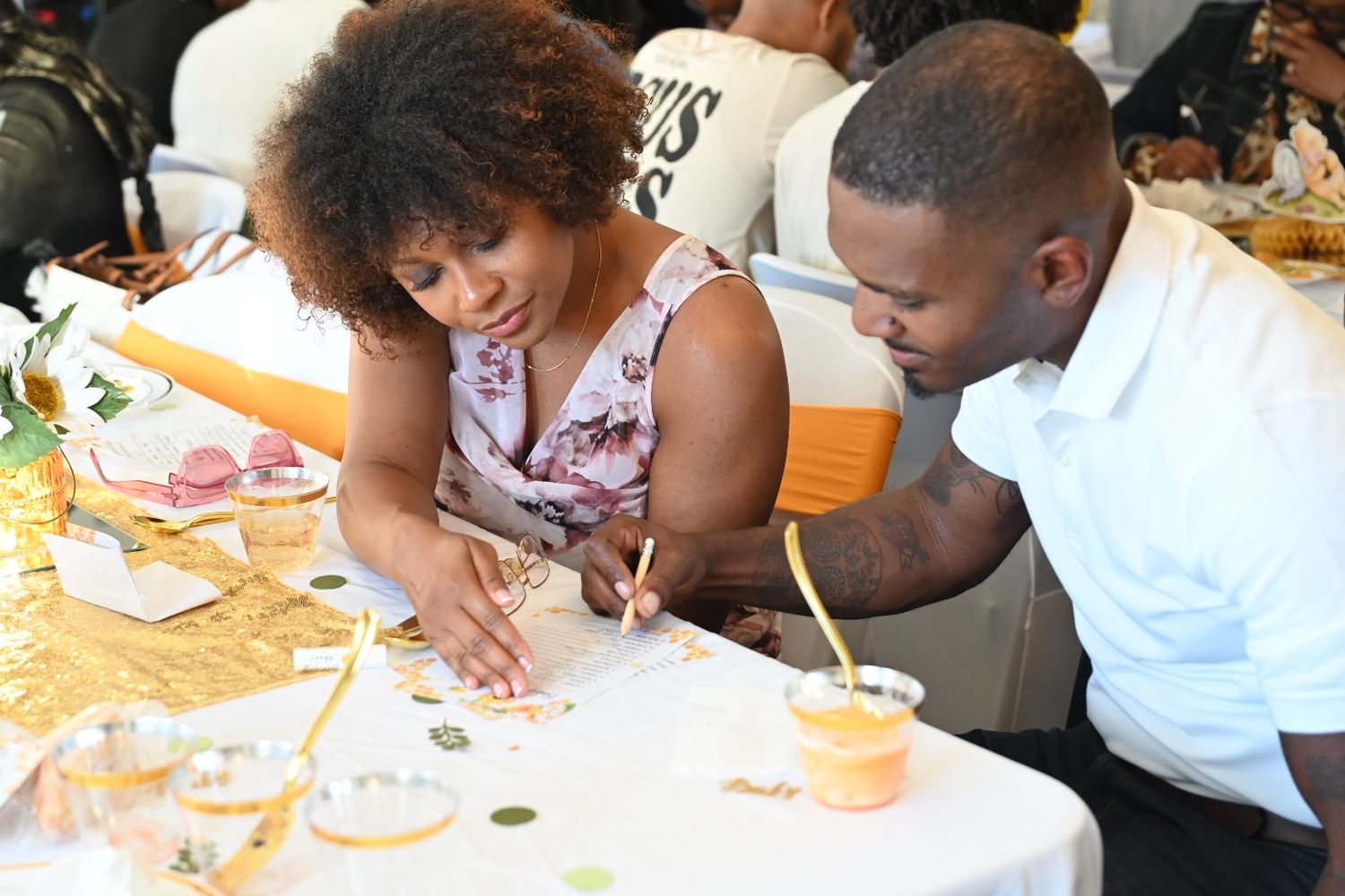 A woman and man collaborate on a craft activity at a decorated baby shower table with gold accents.