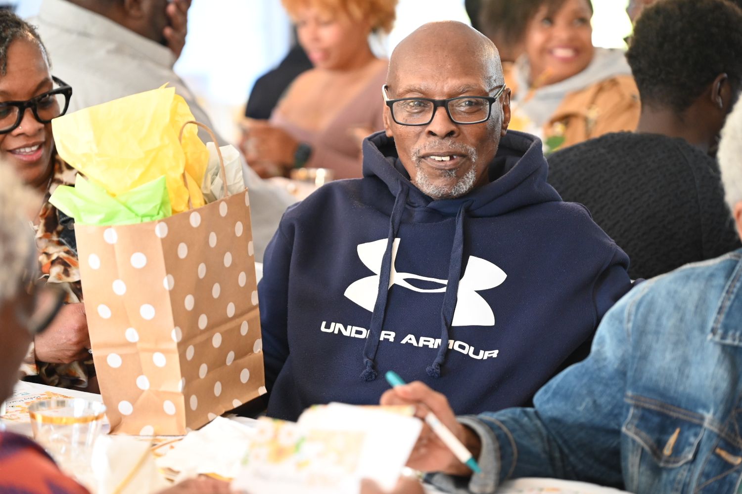 Man in Under Armour hoodie sits at a baby shower table with a polka dot gift bag.