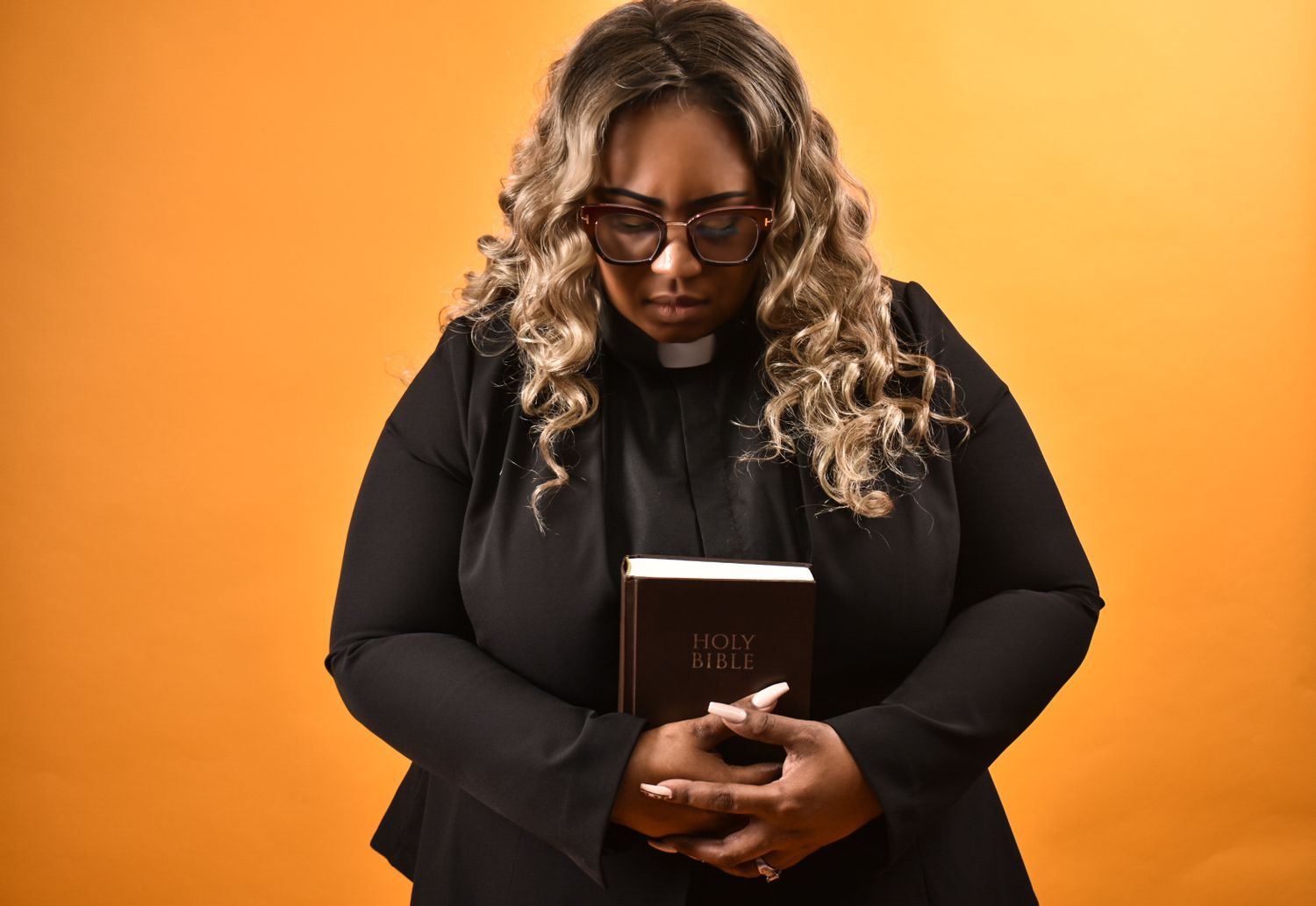 Black female pastor in glasses holding Holy Bible while looking down, wearing black clergy attire.
