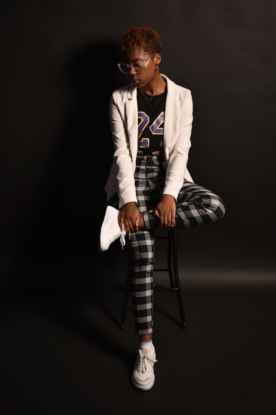 Young woman in white blazer and plaid pants sitting on stool against dark background.