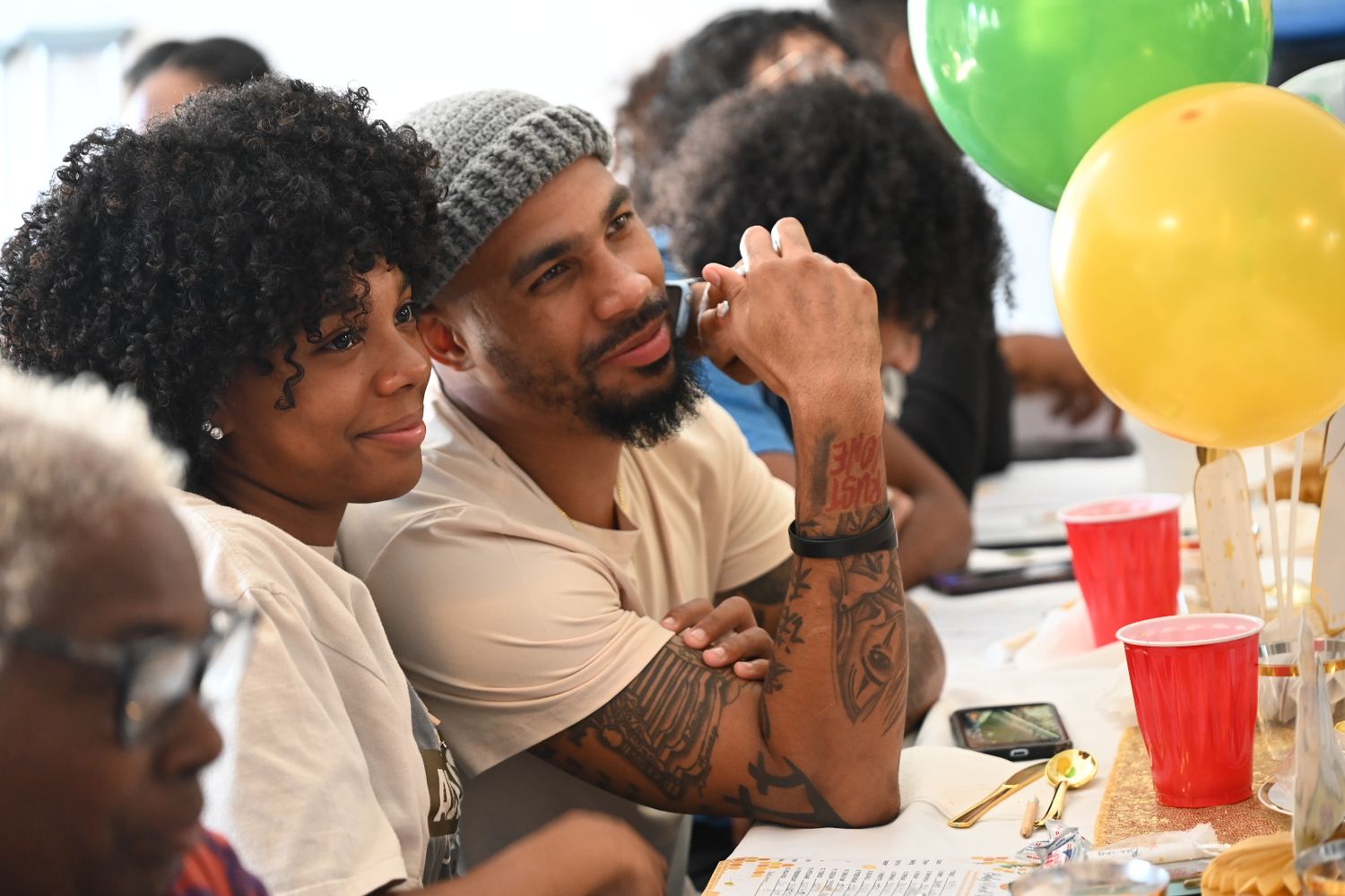 A tattooed man and woman sit together at a colorful party with balloons.