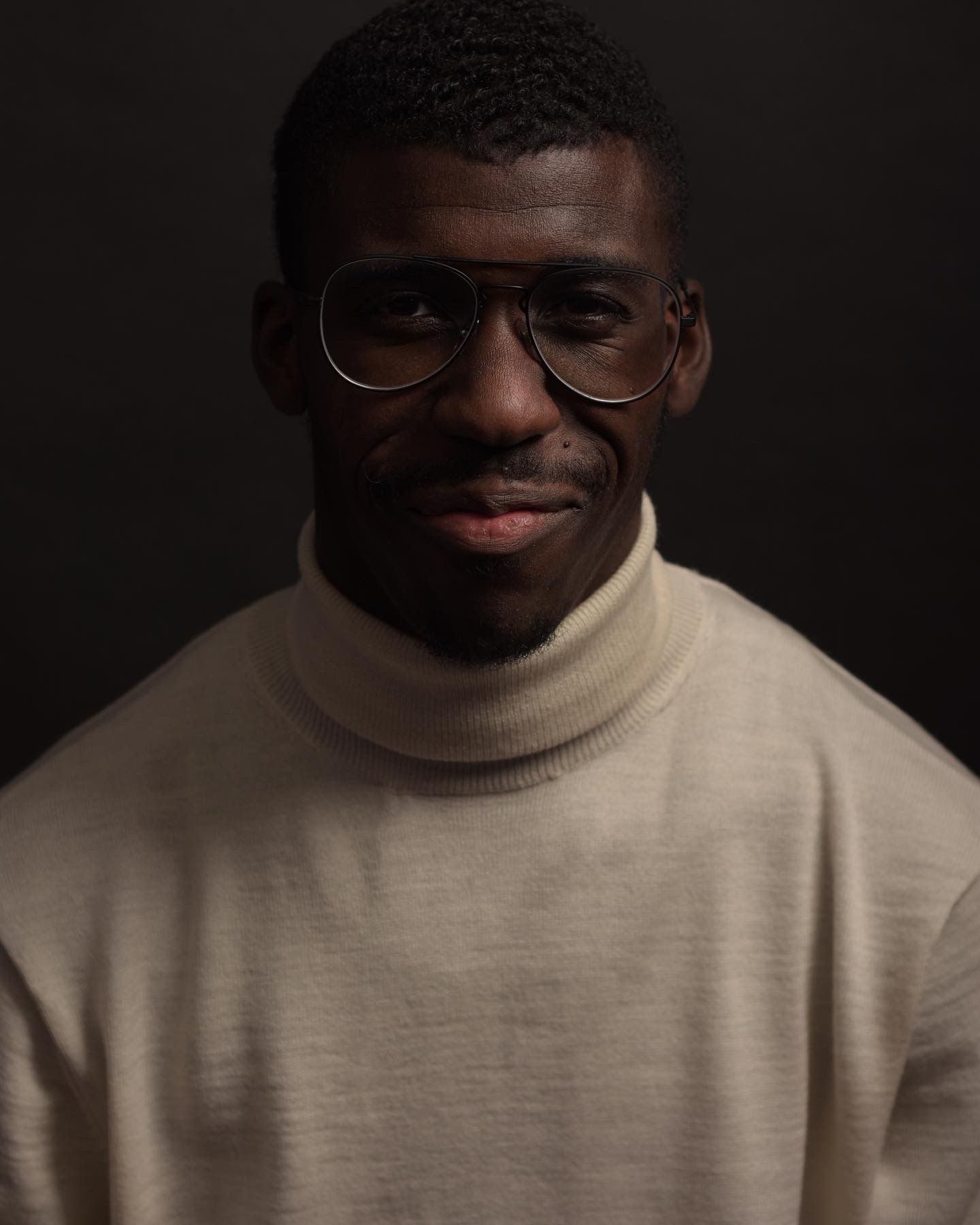 Smiling Black man wearing aviator glasses and a beige turtleneck sweater against a dark background.