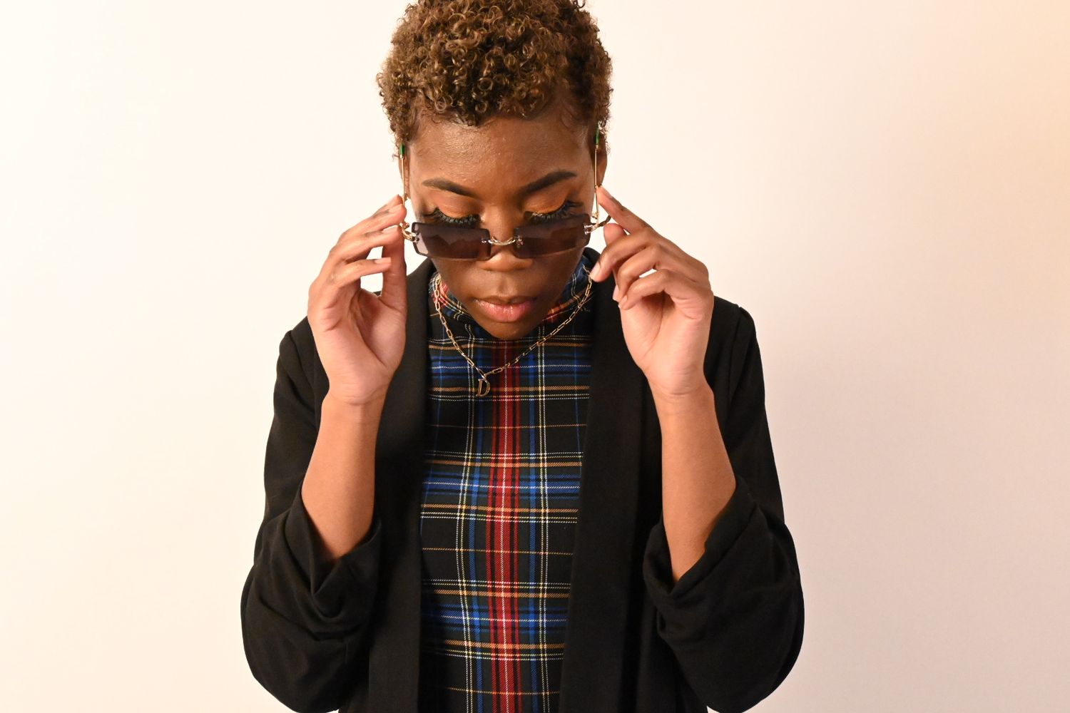 Black person in plaid shirt and blazer adjusts round sunglasses against white background.