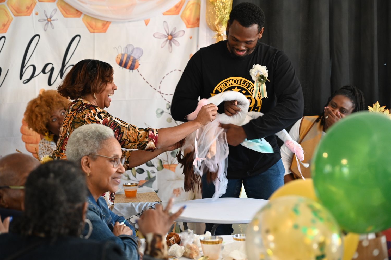 Guests admire baby gifts at a festive bee-themed baby shower with green and gold balloons.