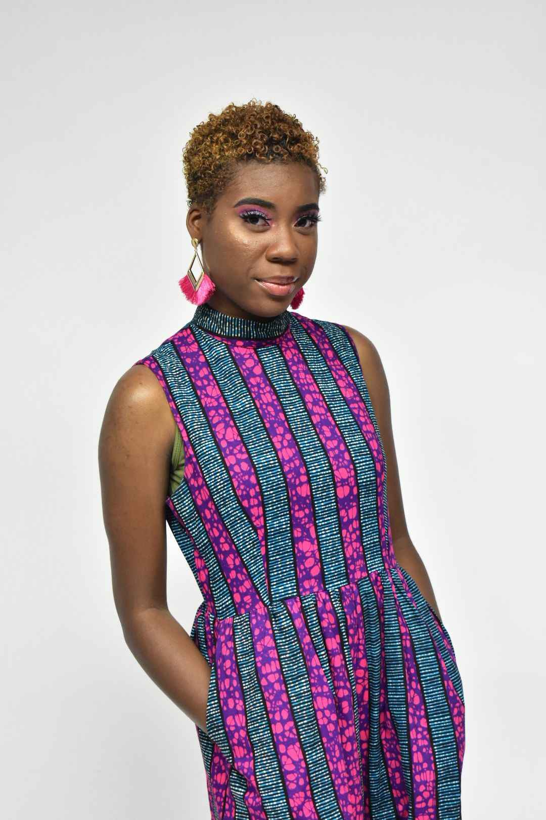 Woman in vibrant African print sleeveless dress with pink tassel earrings on white background.