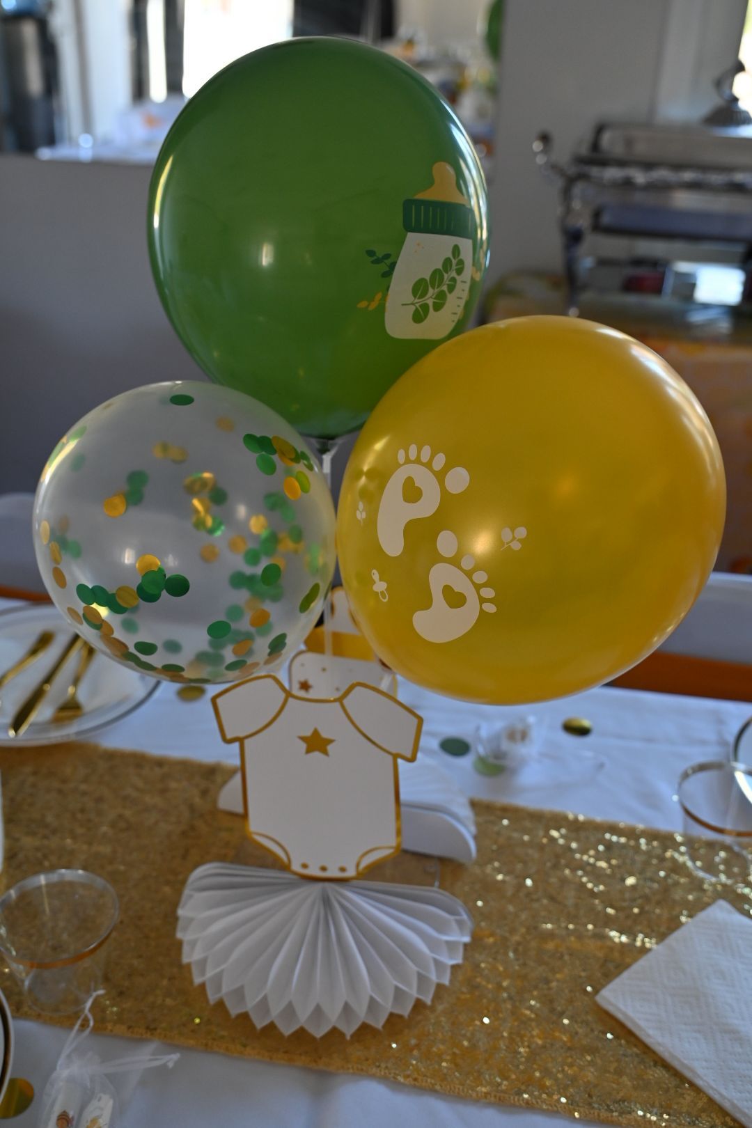 Yellow and green baby shower balloon centerpiece with footprint and bottle balloons on glitter table.