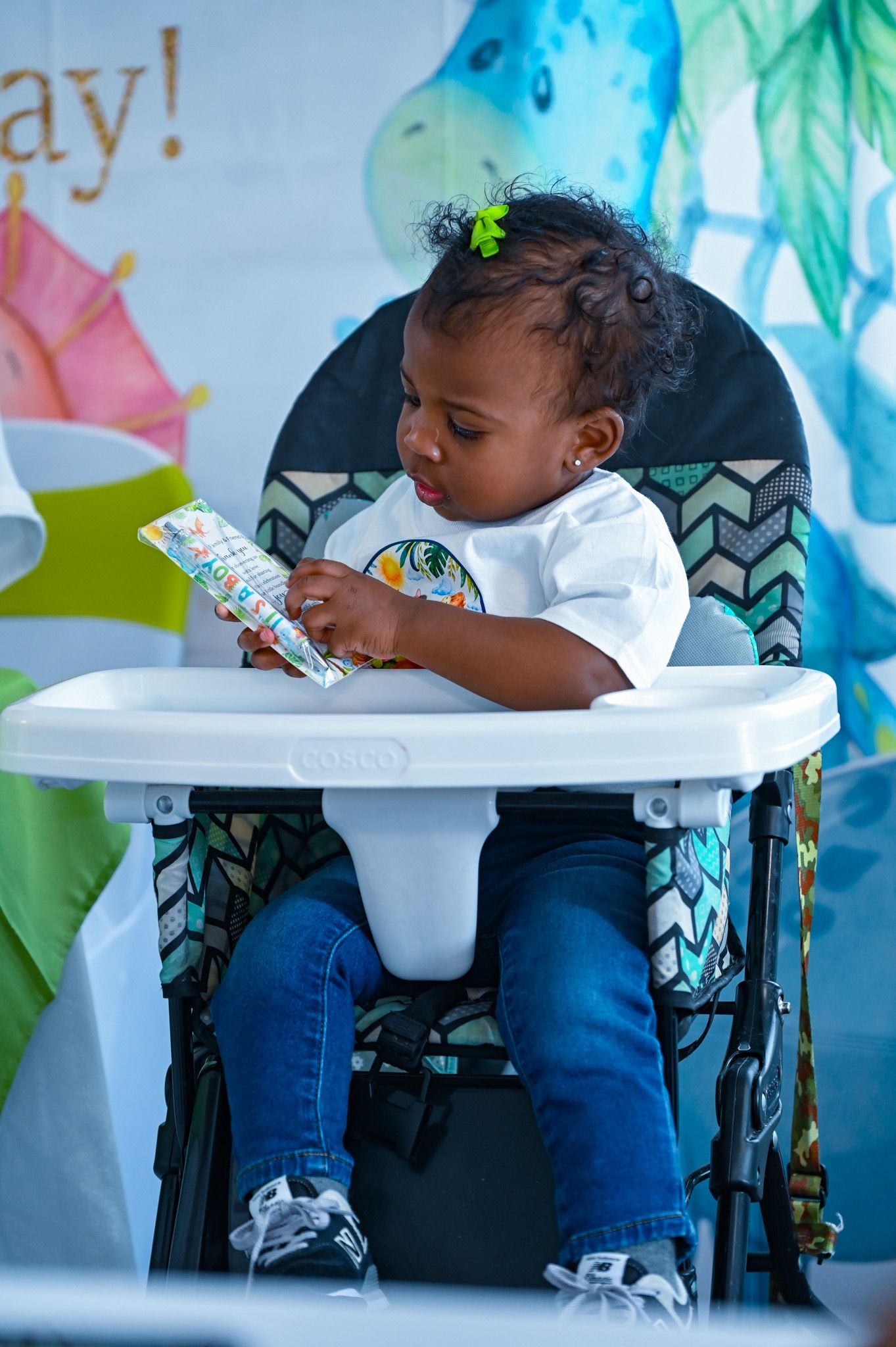 Baby boy in high chair reading a book at a colorful dinosaur-themed birthday party.