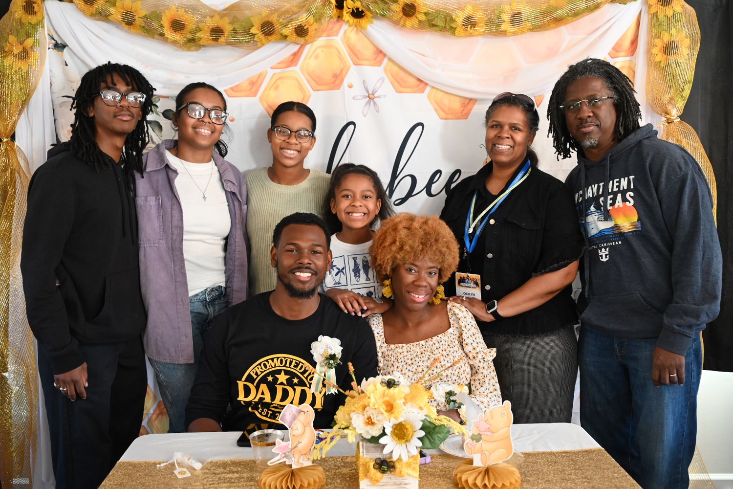 Group of eight people smiling at a baby shower with an oh babee backdrop and sunflower decorations.
