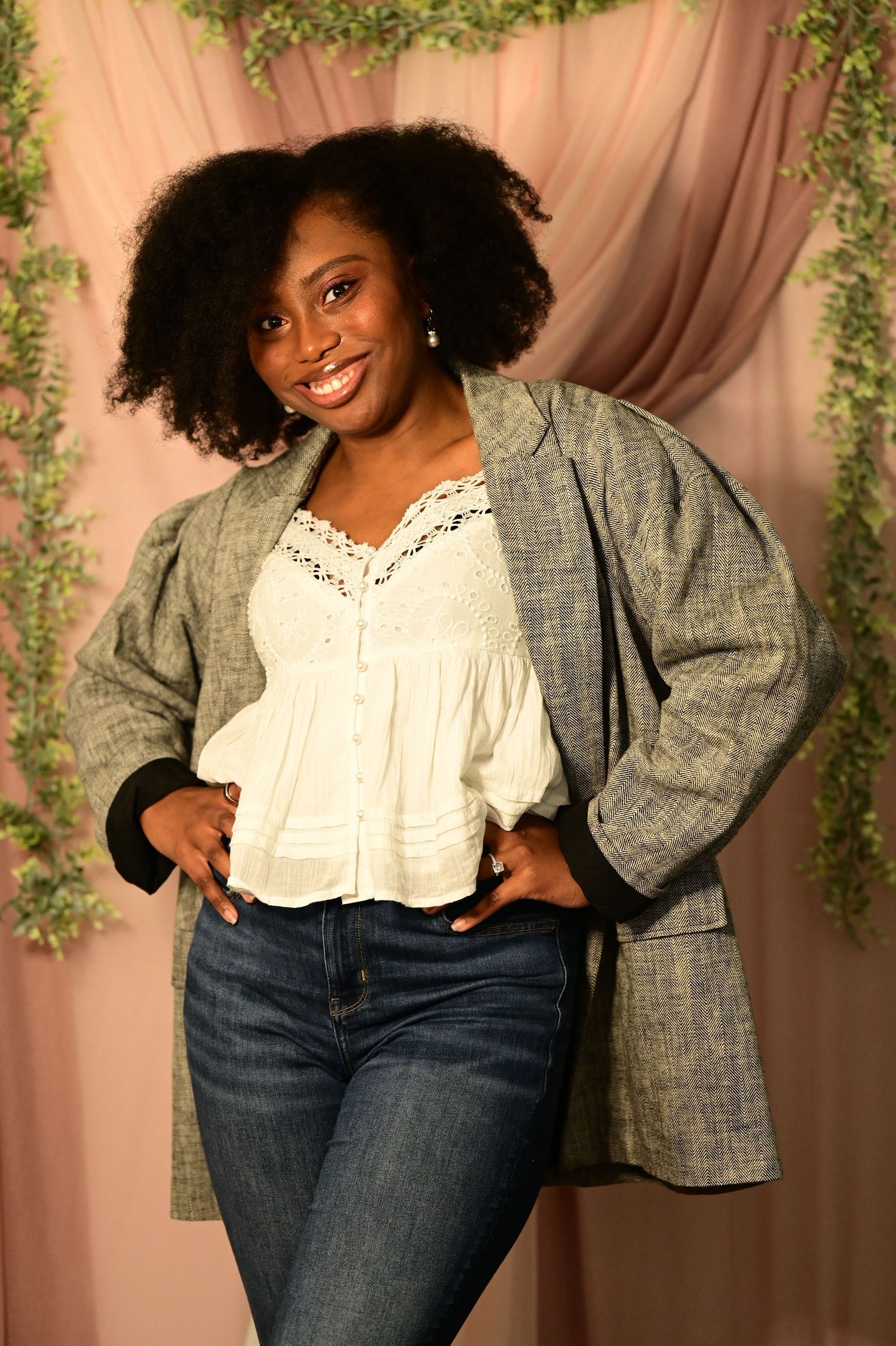 Smiling woman in blazer and jeans posing in front of a pink backdrop with greenery.