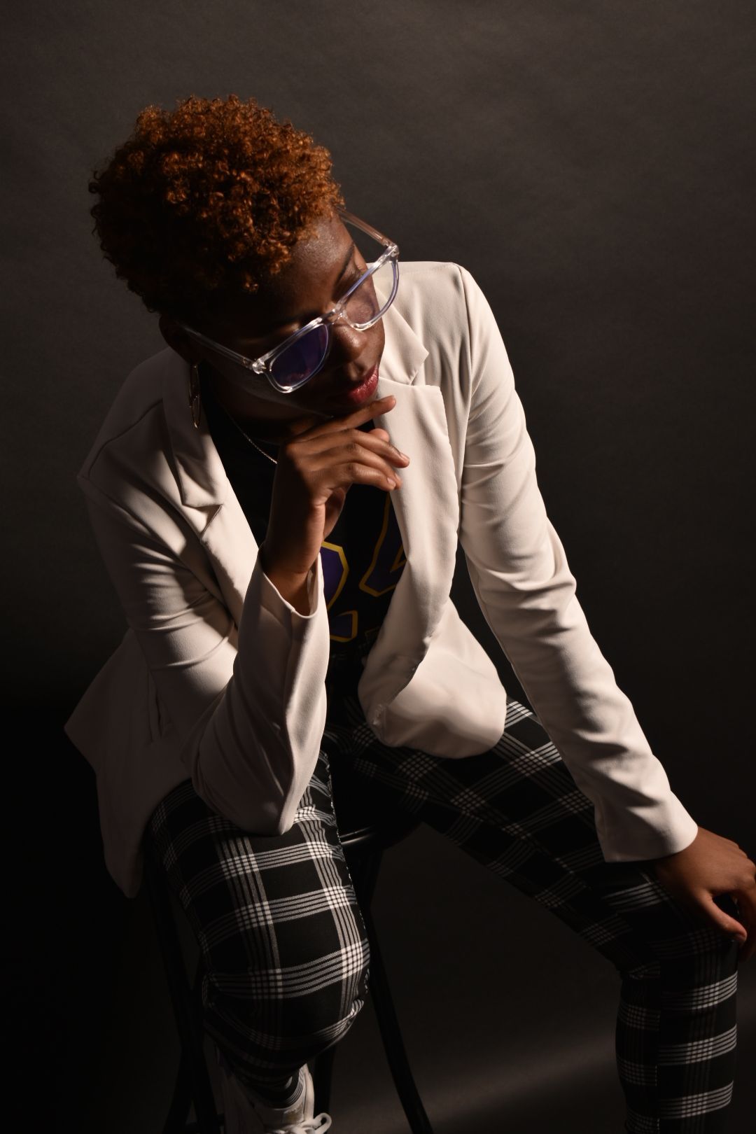 Woman in white blazer and plaid pants posing with purple glasses in dramatic dark studio lighting.