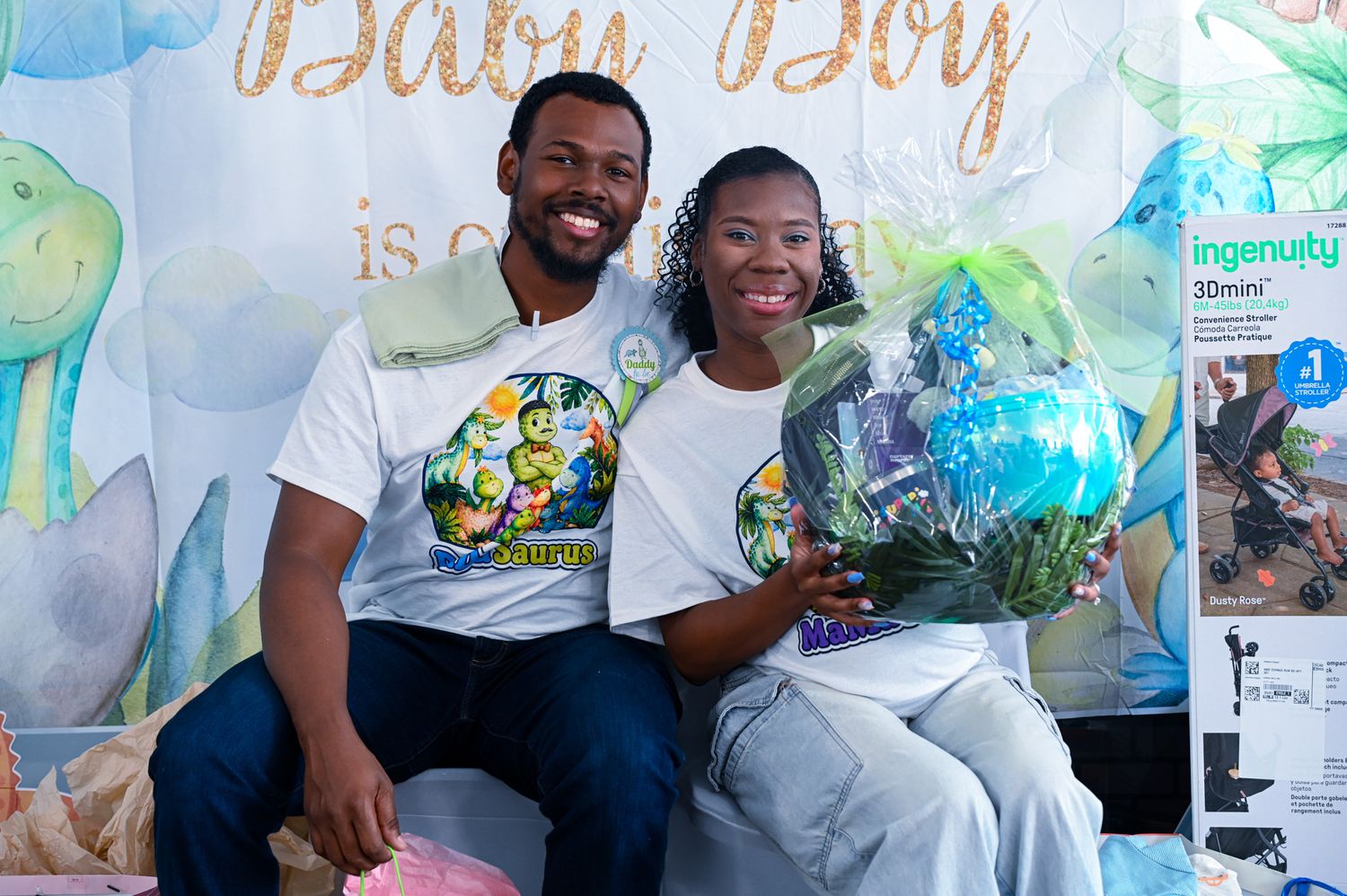 Smiling couple posing with a bouquet and balloon in front of a Baby Boy shower backdrop.