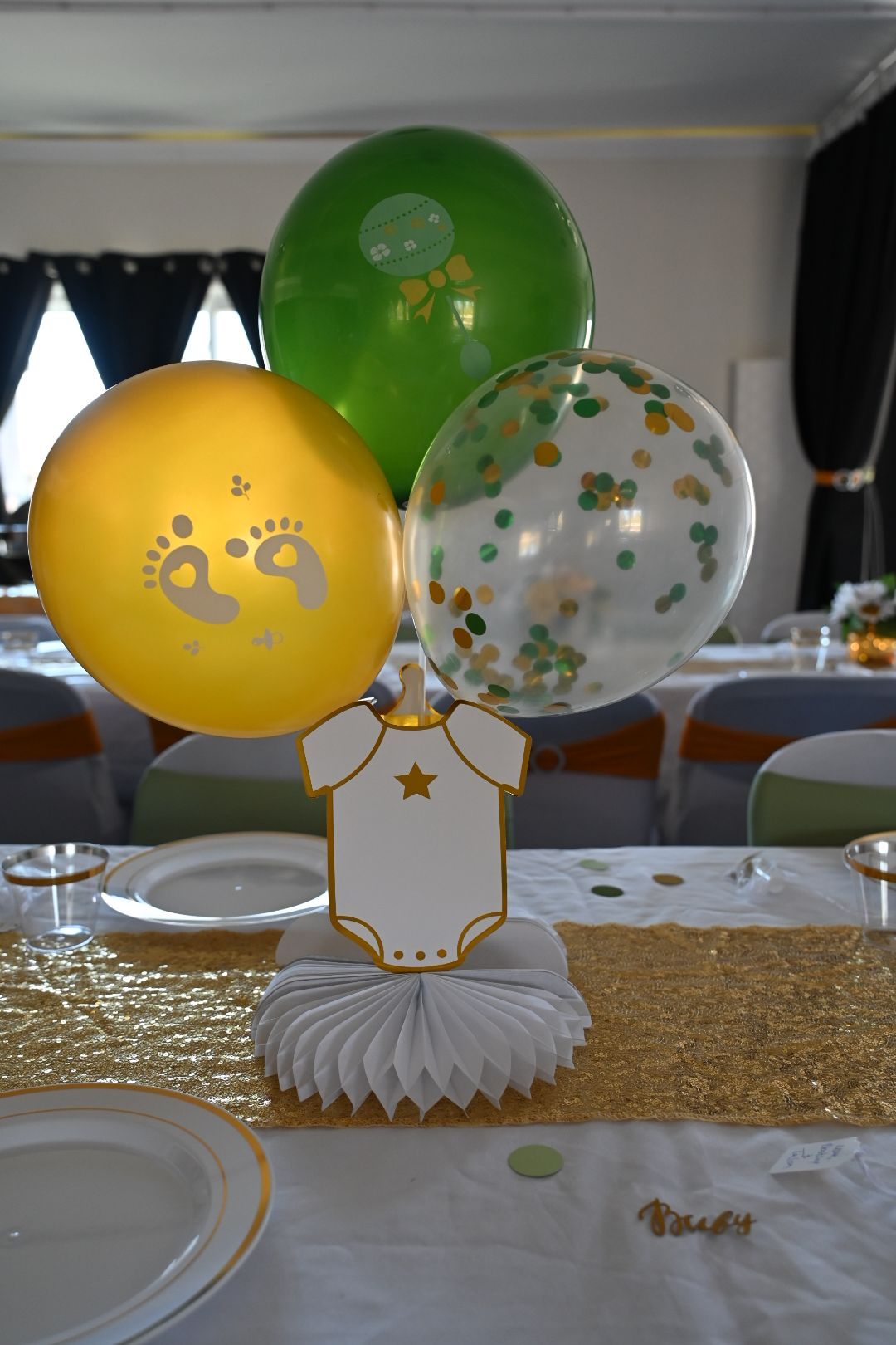 Baby shower balloon centerpiece with gold, green, and confetti balloons above onesie card holder.