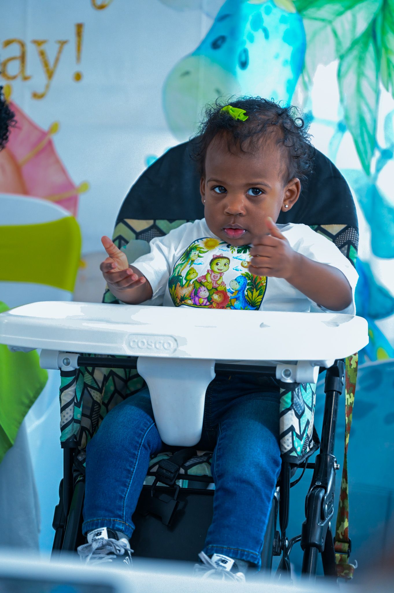Adorable baby boy in high chair giving thumbs up at dinosaur-themed birthday party.