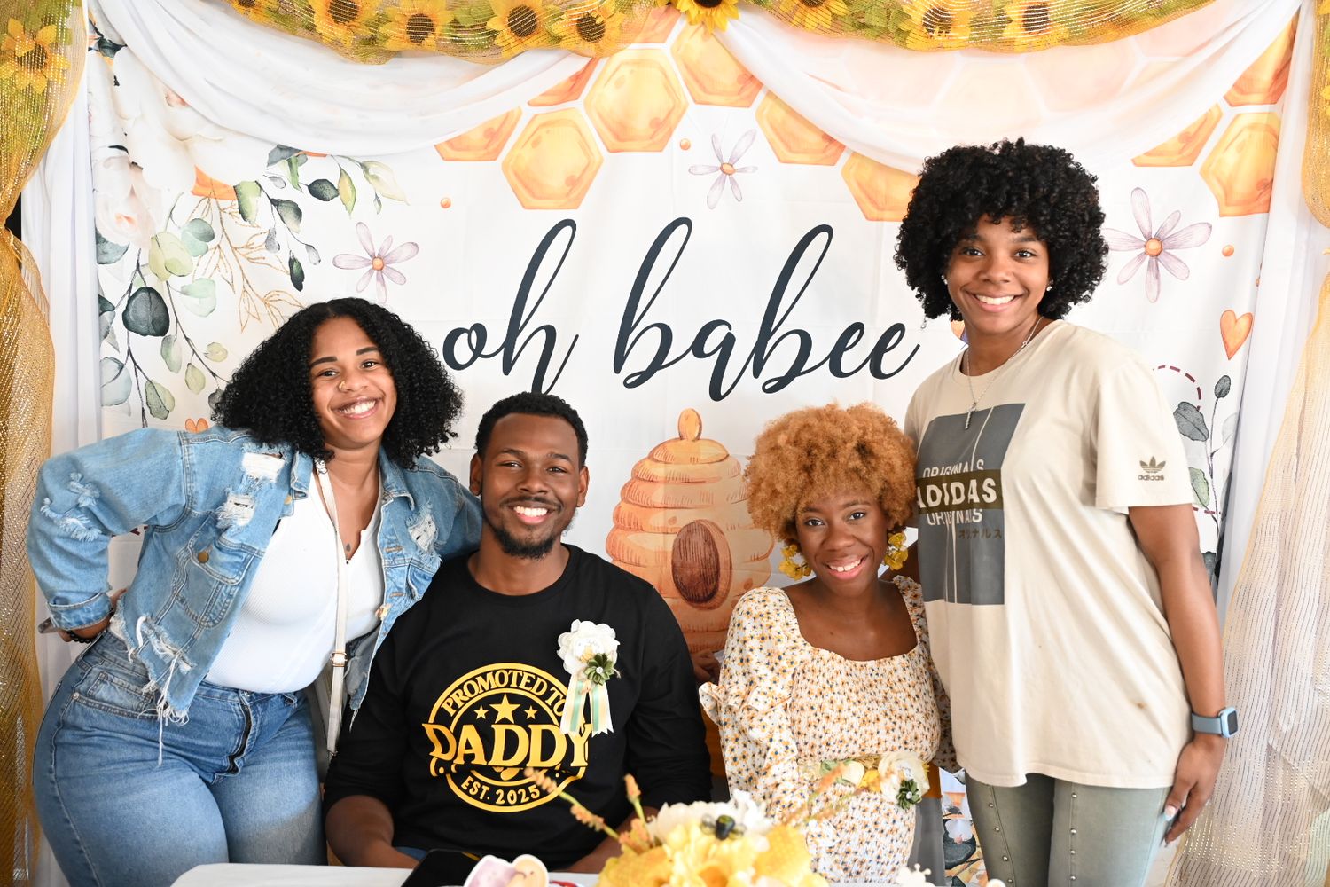 Four people posing happily at a baby shower with an oh babee floral backdrop and sunflowers.