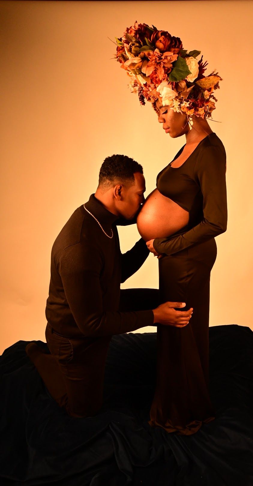 Pregnant Black woman in floral crown stands as partner kneels kissing her bare belly in studio.