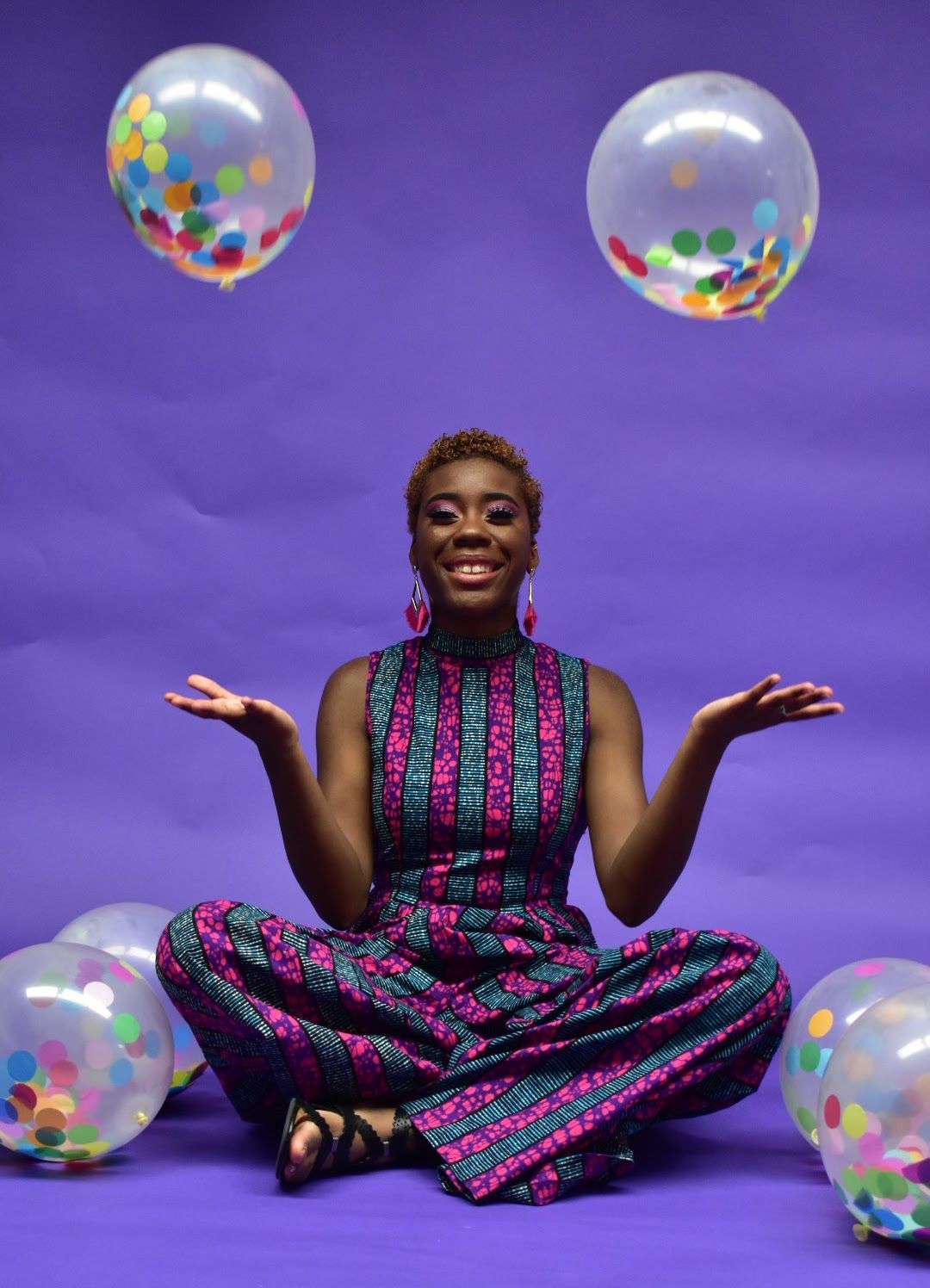 Smiling woman in colorful printed jumpsuit sits cross-legged tossing confetti balloons on purple backdrop.