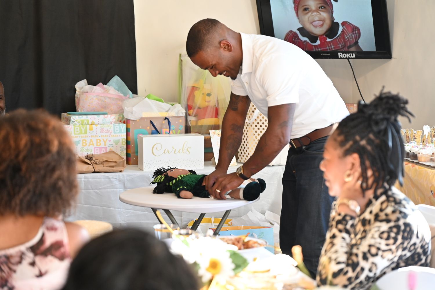 A man in white shirt handles a stuffed toy at a baby shower gift table with diapers and presents.