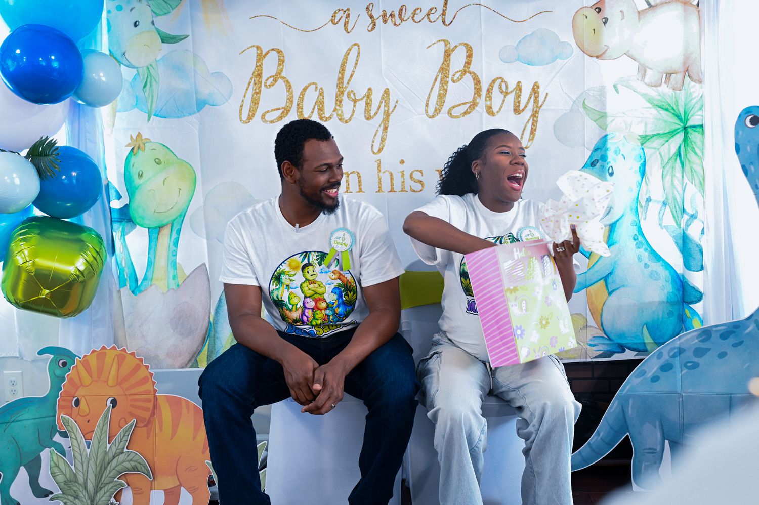 Couple laughing while opening gifts at a Baby Boy themed baby shower with dinosaur decorations.