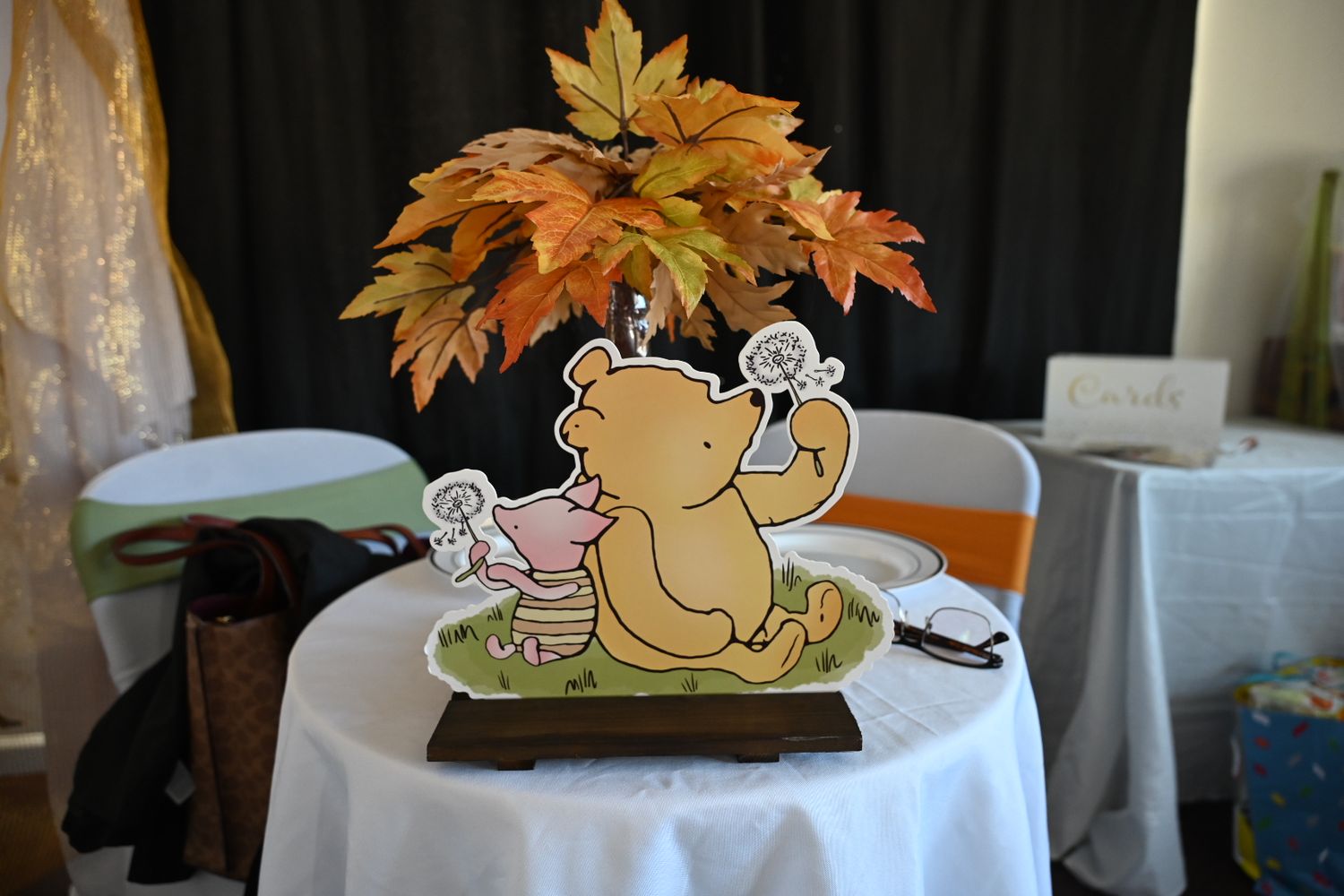 Classic Winnie the Pooh and Piglet centerpiece with autumn leaves on a white tablecloth at baby shower.