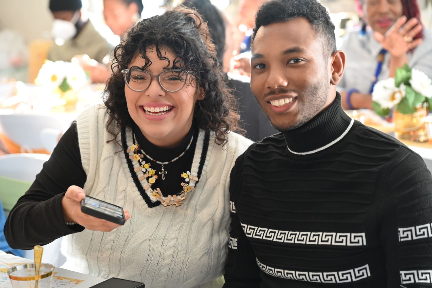 Two smiling people pose together at a baby shower event, woman holding a phone.