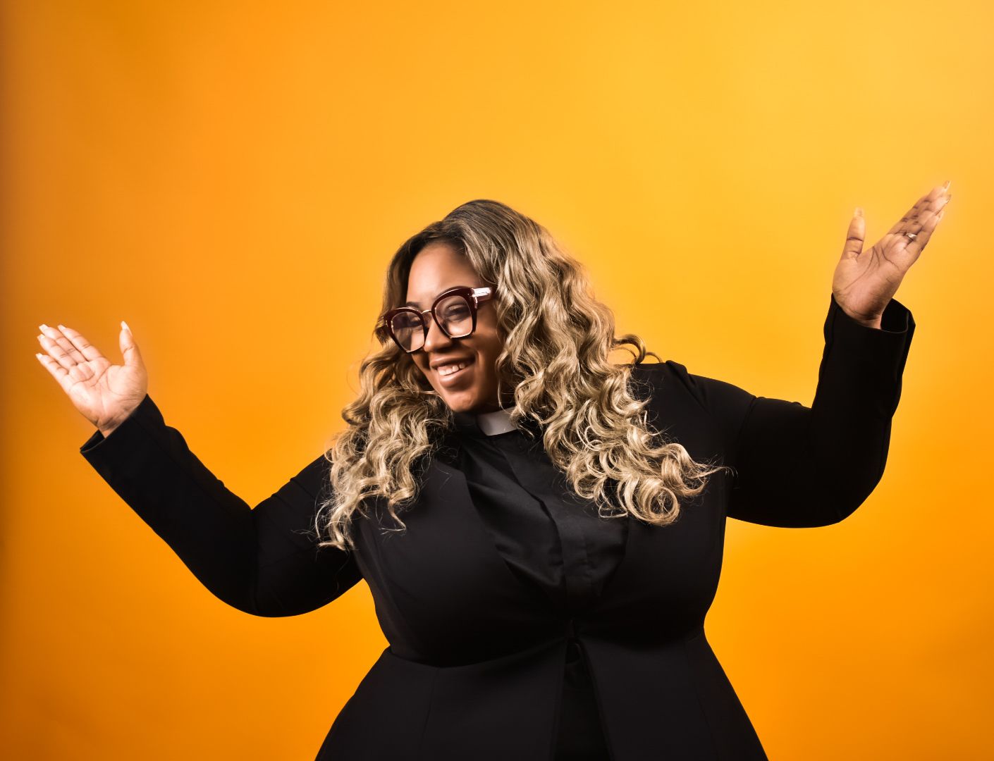 Joyful plus-size Black woman in black outfit and glasses posing against a bright orange background.