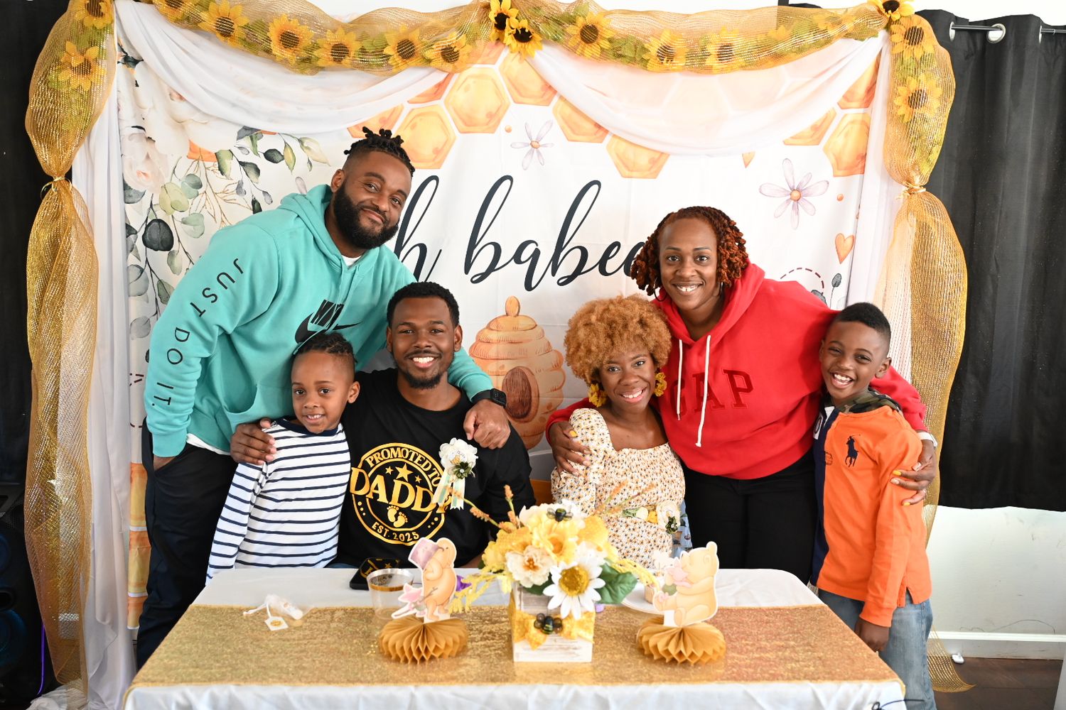A family group of seven poses at a bee-themed baby shower with sunflowers and festive decorations.