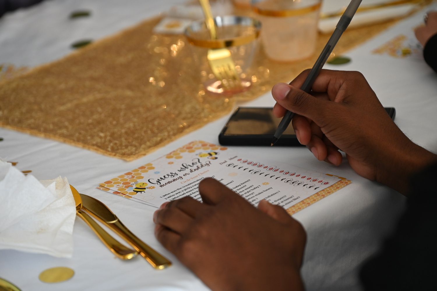 A guest fills out a baby shower game card on a gold and white decorated table with golden utensils.