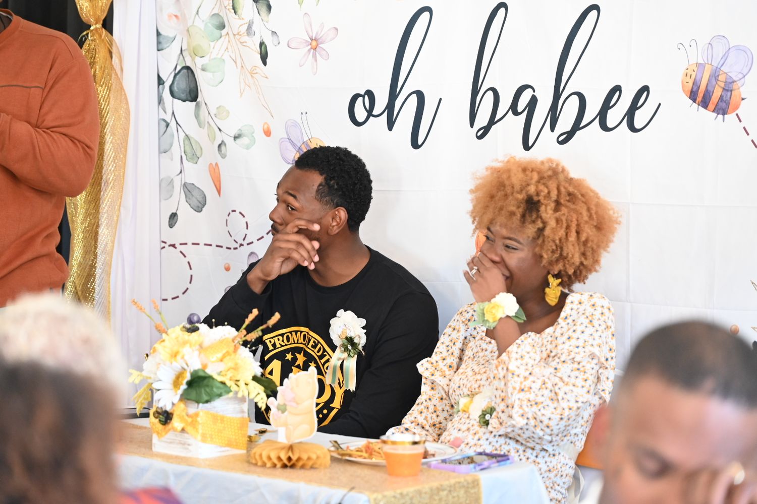 A couple smiles and chats at a bee-themed baby shower with yellow floral centerpieces.
