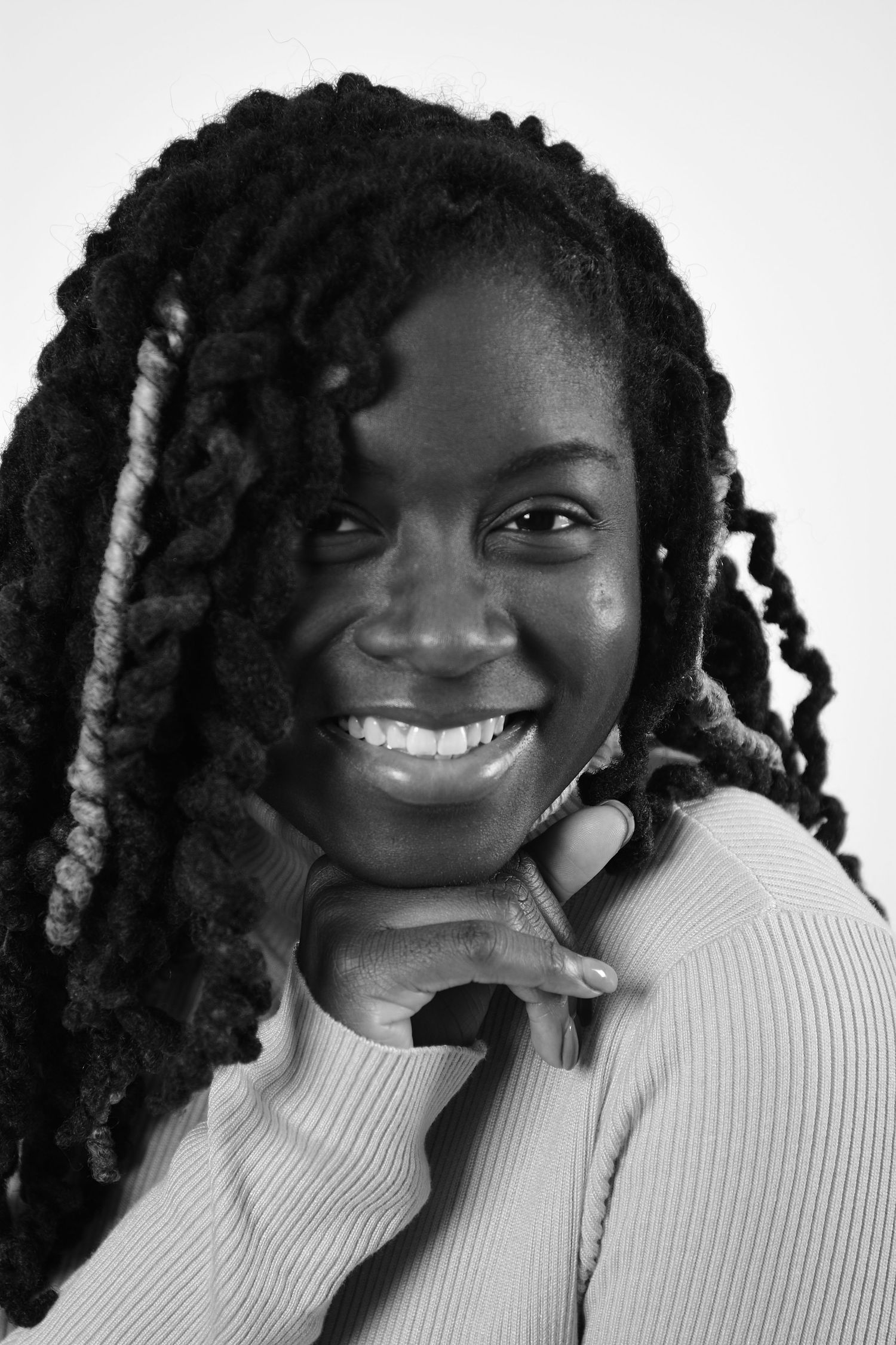 Black and white close-up portrait of a smiling woman with locs resting chin on hands.