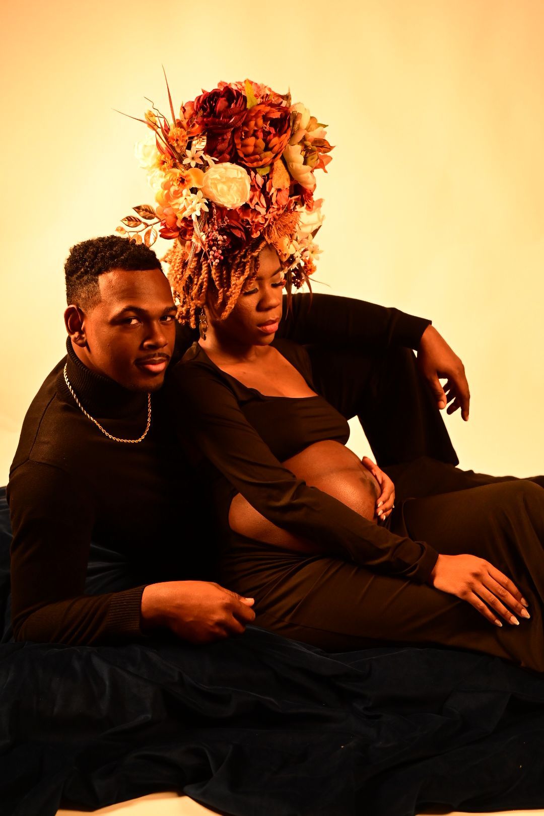 Couple sitting together in maternity photoshoot, woman wearing elaborate floral crown against warm amber background.
