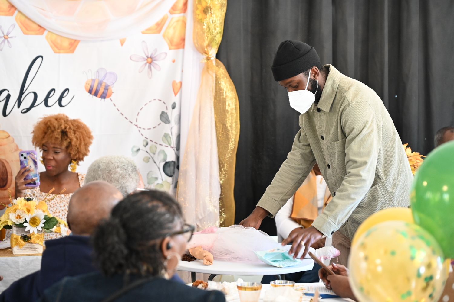 A masked man in olive jacket opens gifts at a bee-themed baby shower with balloon decorations.