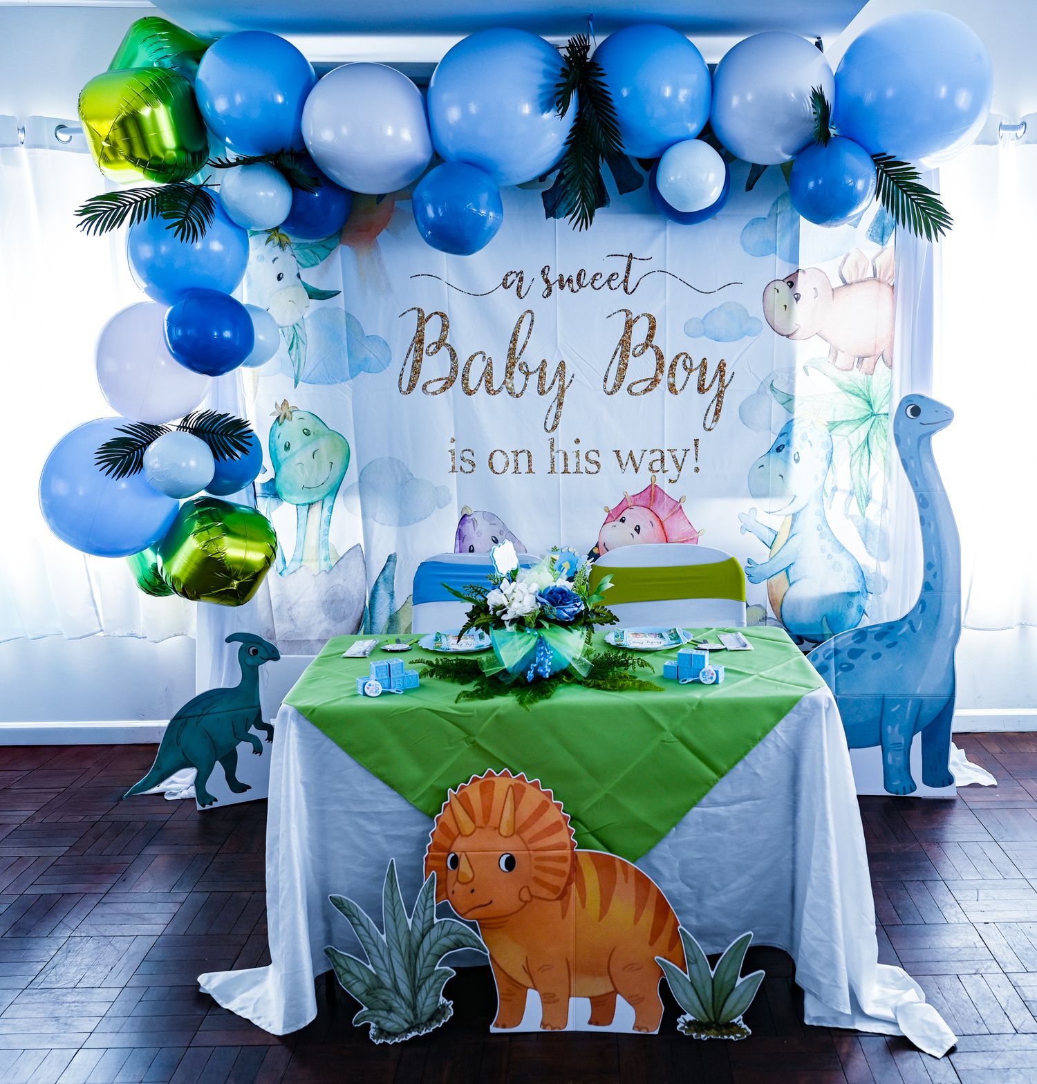 Sweet baby boy dinosaur shower backdrop with blue balloon arch and decorated table.