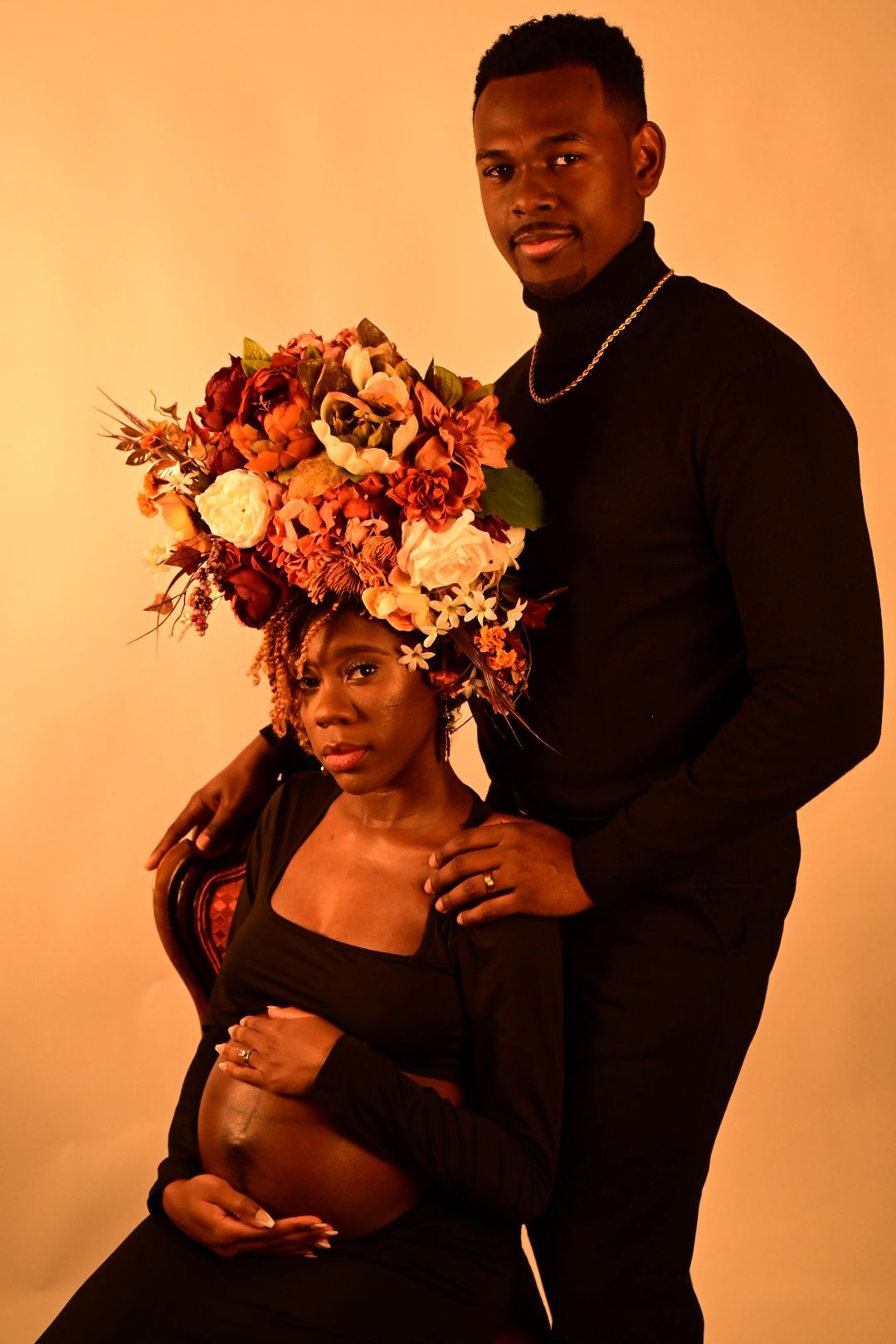 Maternity portrait of couple in black, pregnant woman seated with floral crown while partner stands behind her.