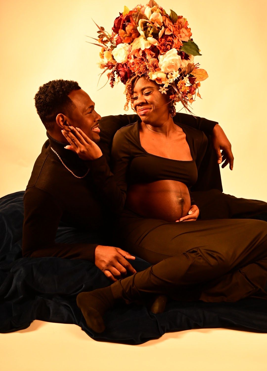 Happy expecting couple sharing a laugh during maternity photoshoot with floral crown and warm amber lighting.