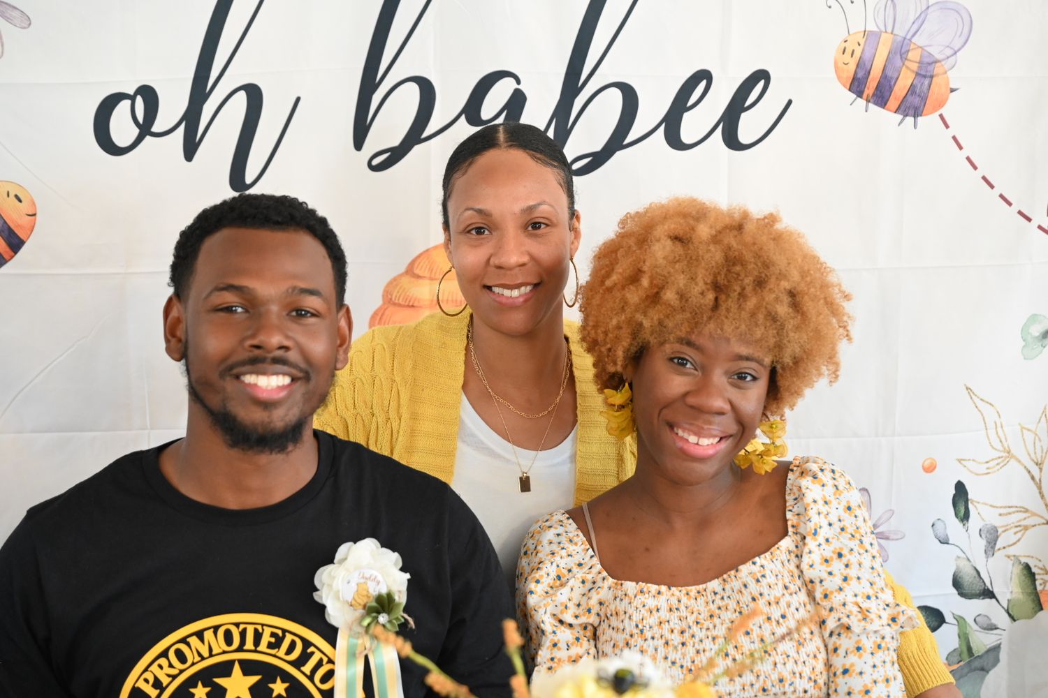 Three adults smiling together at a bee-themed baby shower with oh babee banner behind them.
