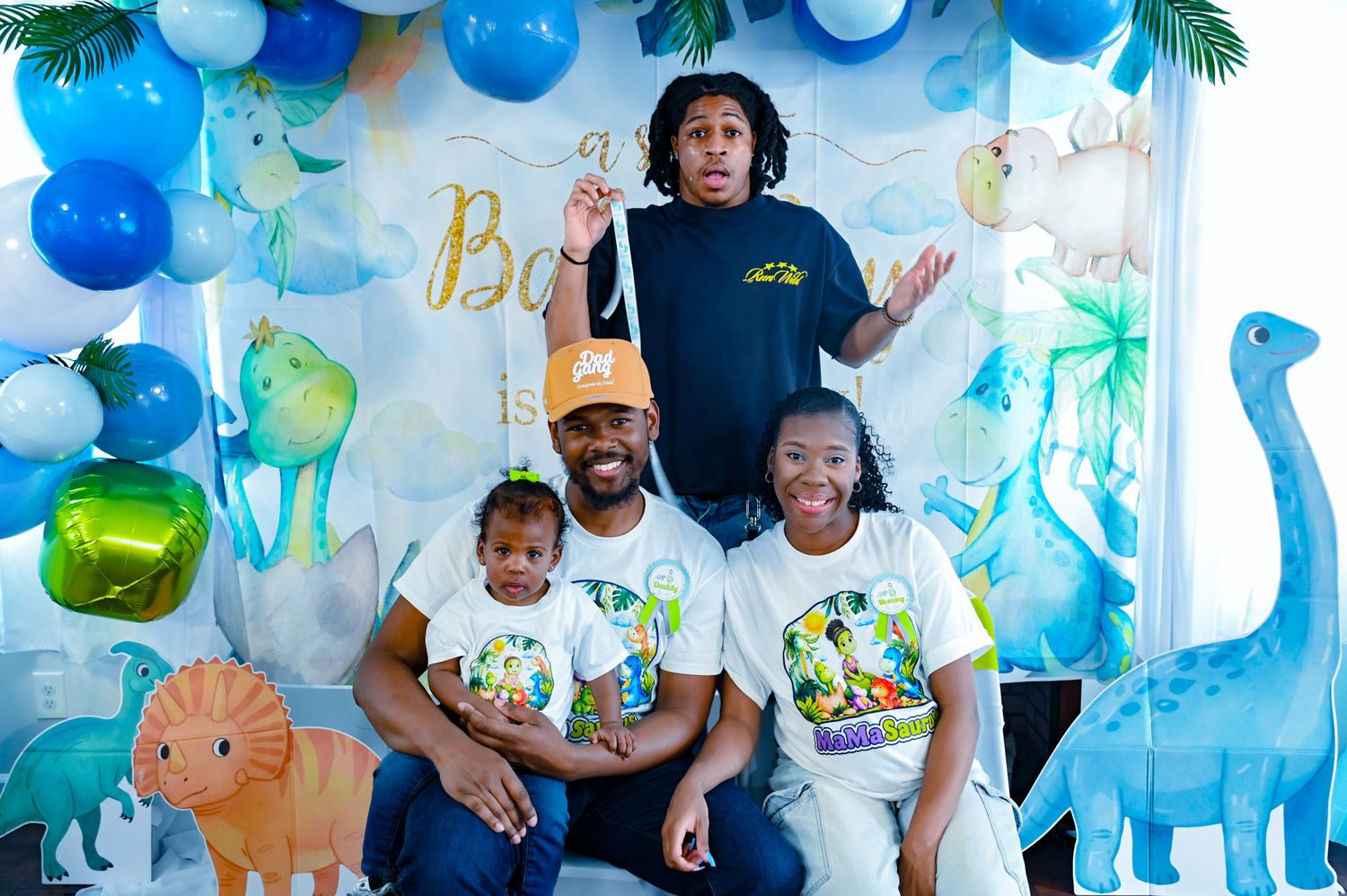 Family of four with teen at a Baby Boy dinosaur birthday party balloon backdrop.