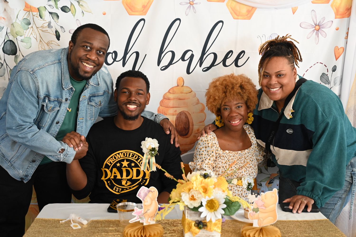Four people smiling at a baby shower with an oh babee backdrop and floral decorations.