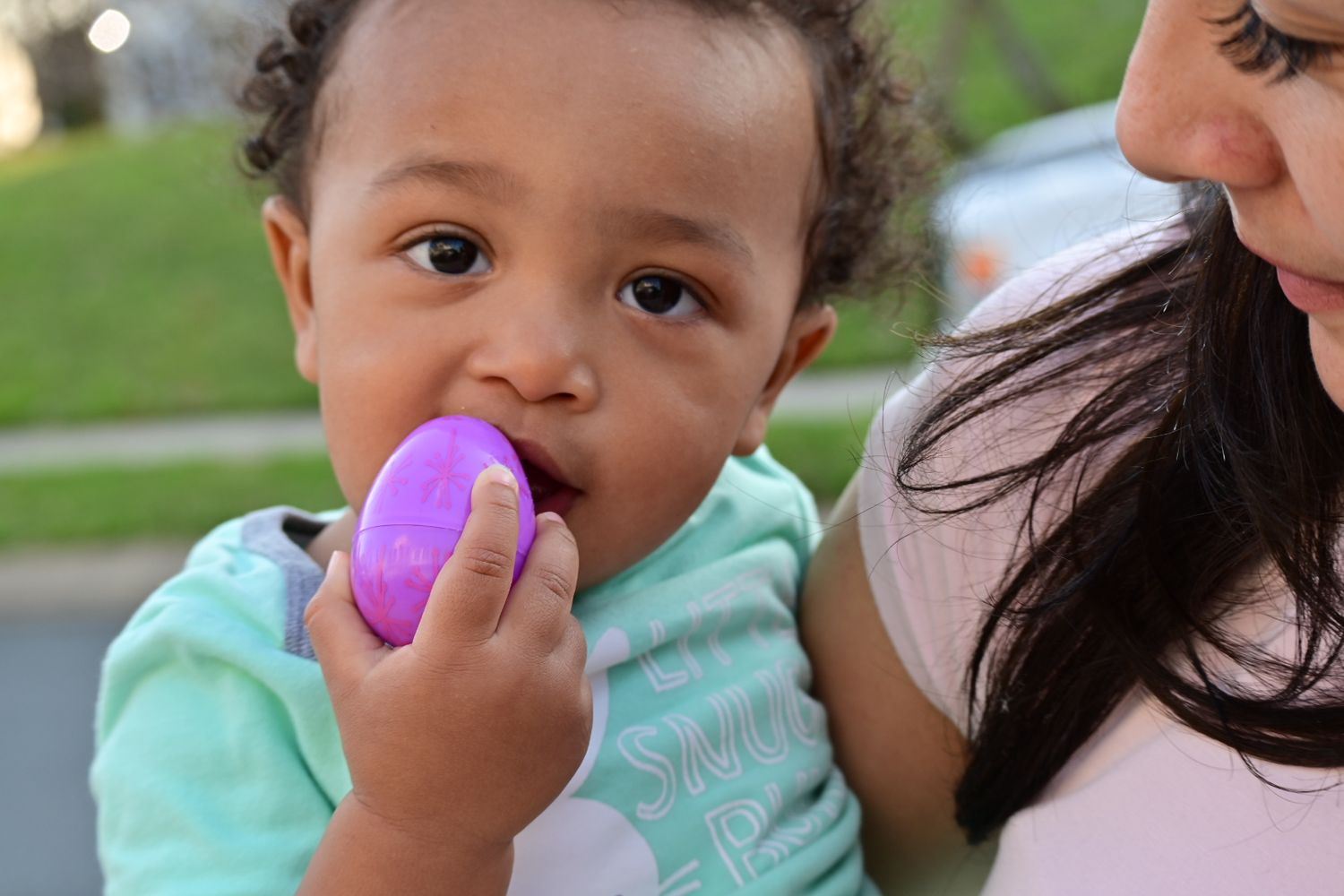 Close-up of a baby chewing on a purple Easter egg while being held outdoors.