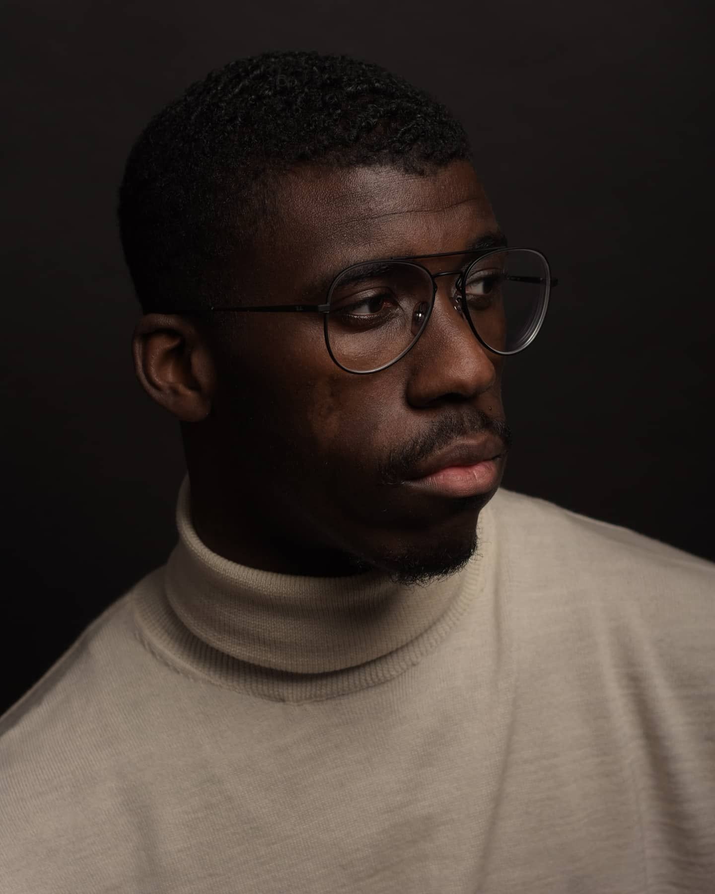 Close-up portrait of a Black man wearing aviator glasses and a beige turtleneck sweater, looking away.