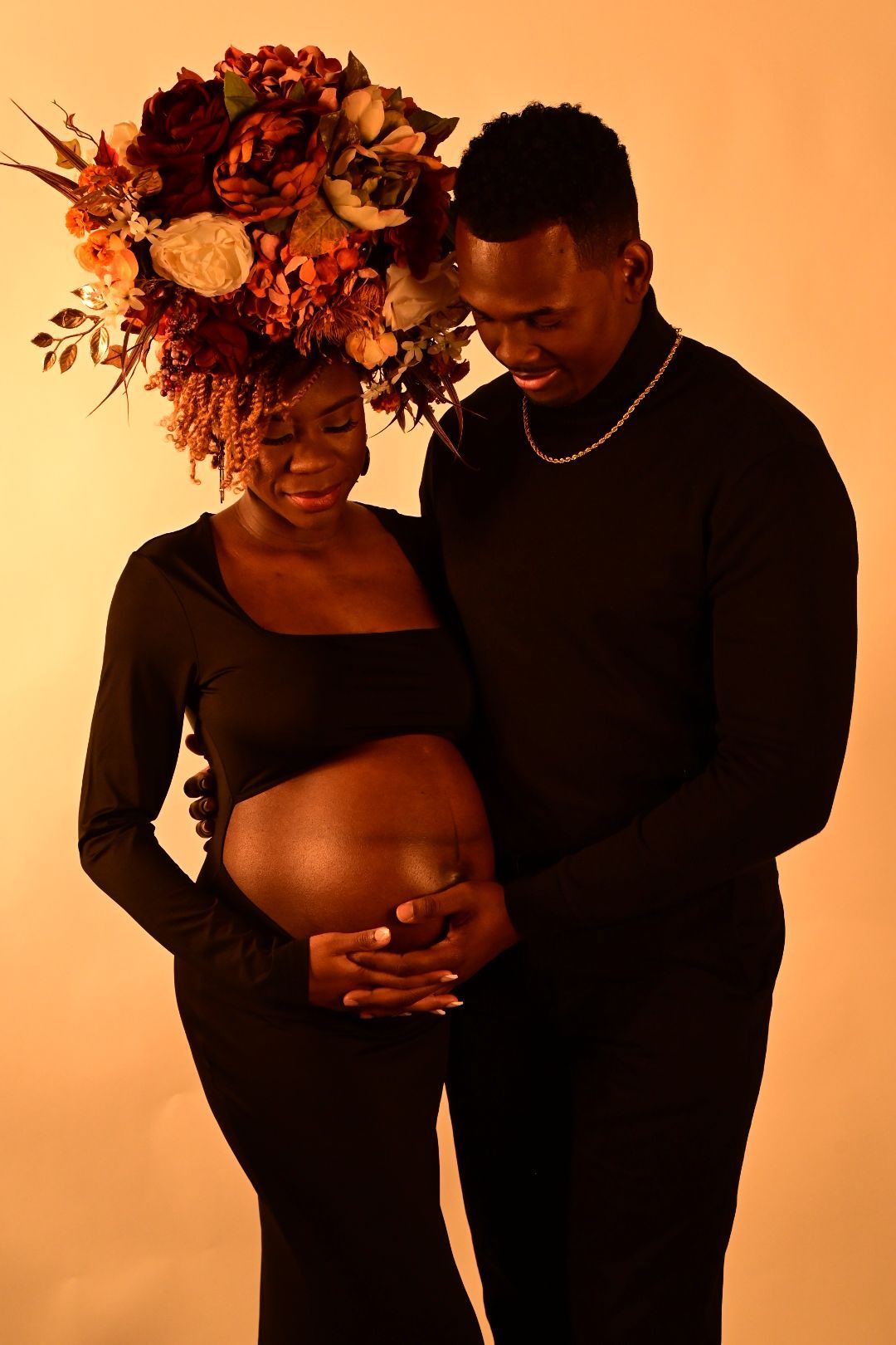 Expecting couple in black outfits pose for maternity shoot, woman wearing stunning floral crown on warm background.